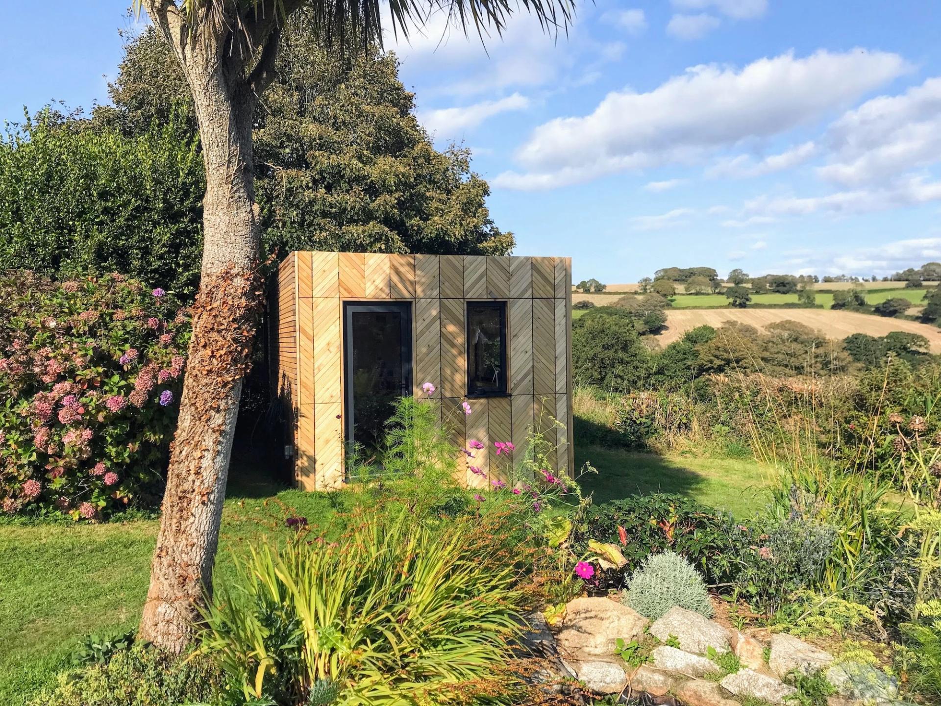 A small sustainable wooden garden shed with a glass door stands among lush greenery, flowering plants, and a palm tree, overlooking rolling fields under a partly cloudy sky.