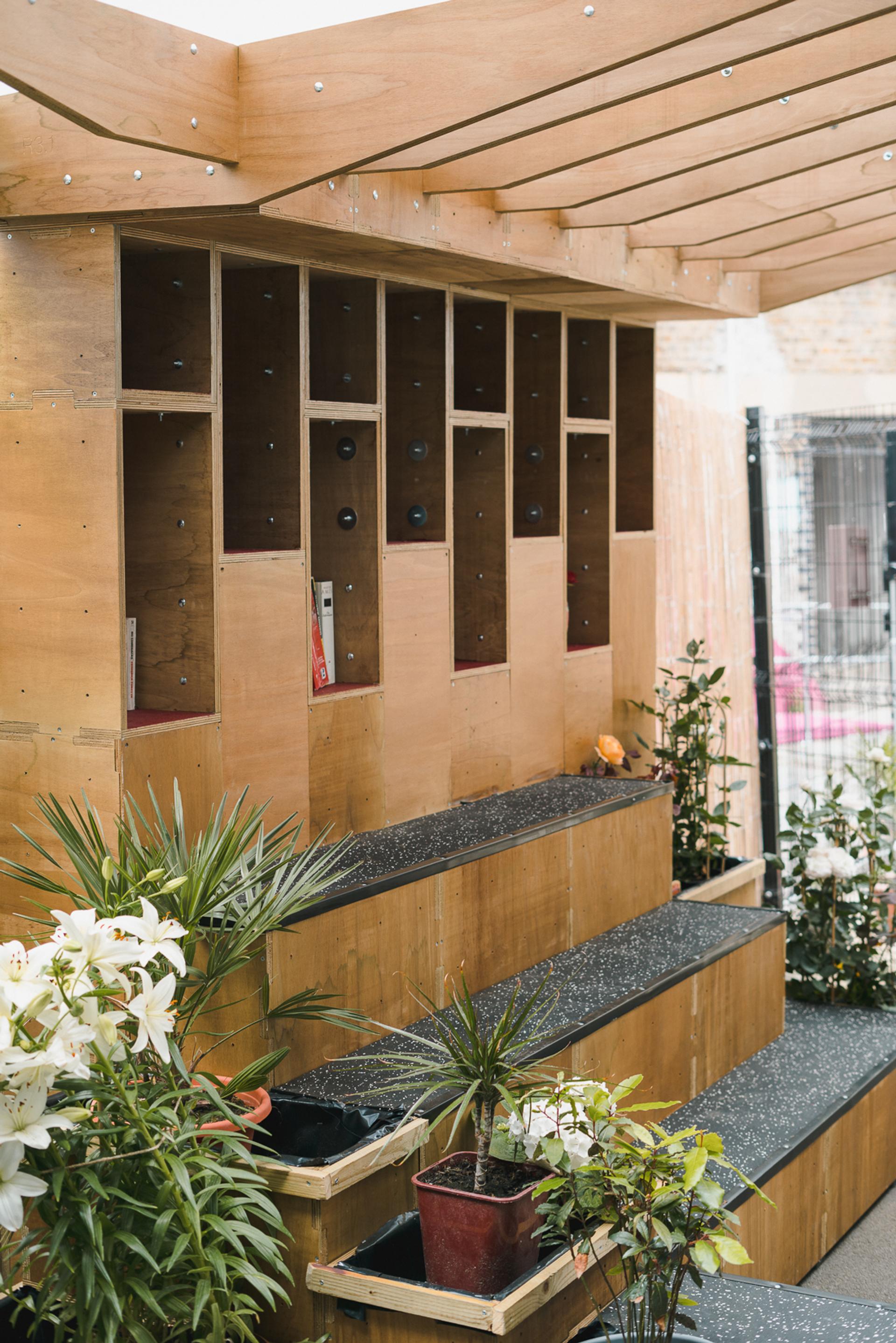 Wooden tiered seating with built-in cubby holes, some containing bocce balls. Flowering plants in pots line the steps and lower level, under a partially covered roof—all crafted from sustainably sourced materials.