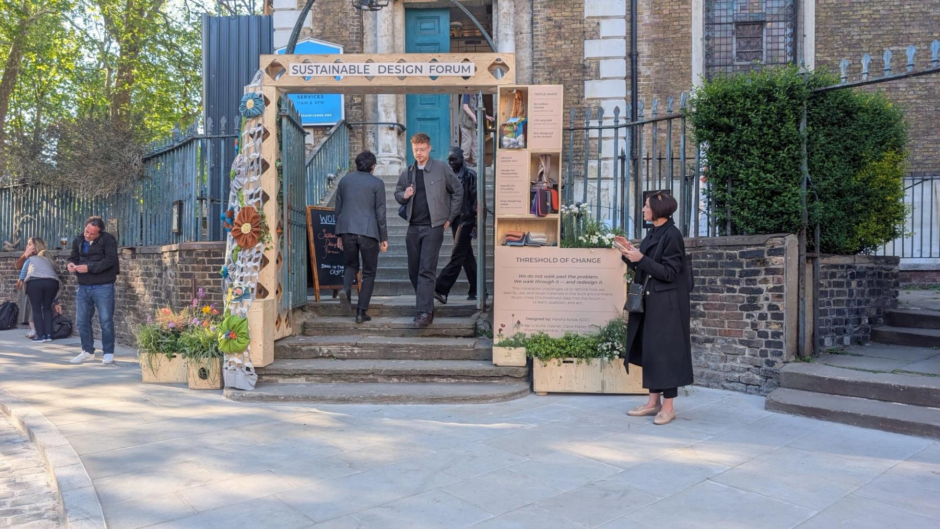 People enter a building through a decorated wooden arch labelled Sustainable Design Forum, part of the Threshold of Change installation. Informational boards stand nearby as a woman in black reads one amid trees and brick walls.