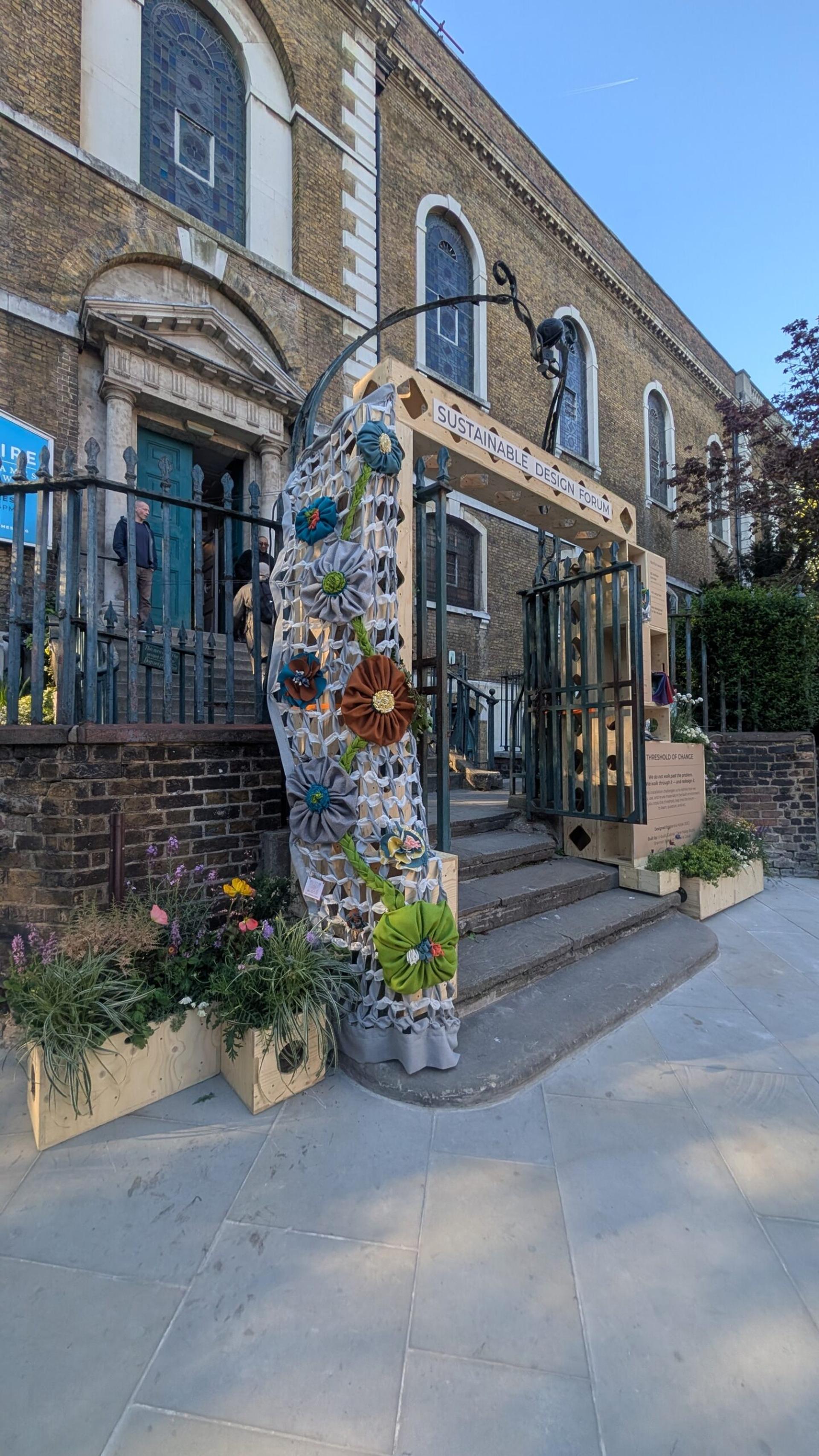 A church entrance decorated with colourful flowers and a sign reading SUSTAINABLE DESIGN marks an installation, symbolising the Threshold of Change. Planters and greenery line the brick exterior with arched windows and a gated doorway.