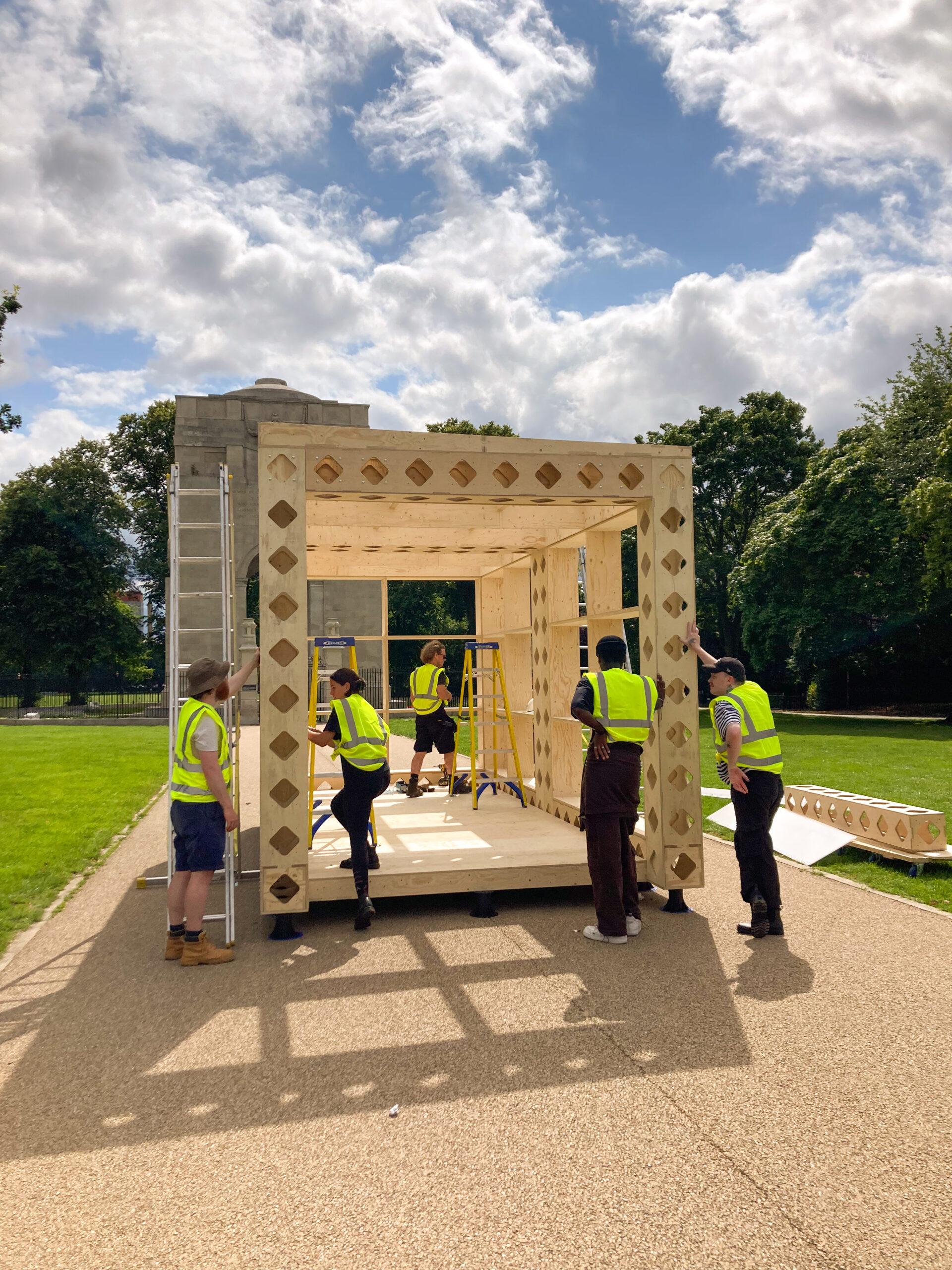Four people in yellow safety vests assemble a sustainable timber construction outdoors on a sunny day, with scattered clouds in the sky and green trees in the background.