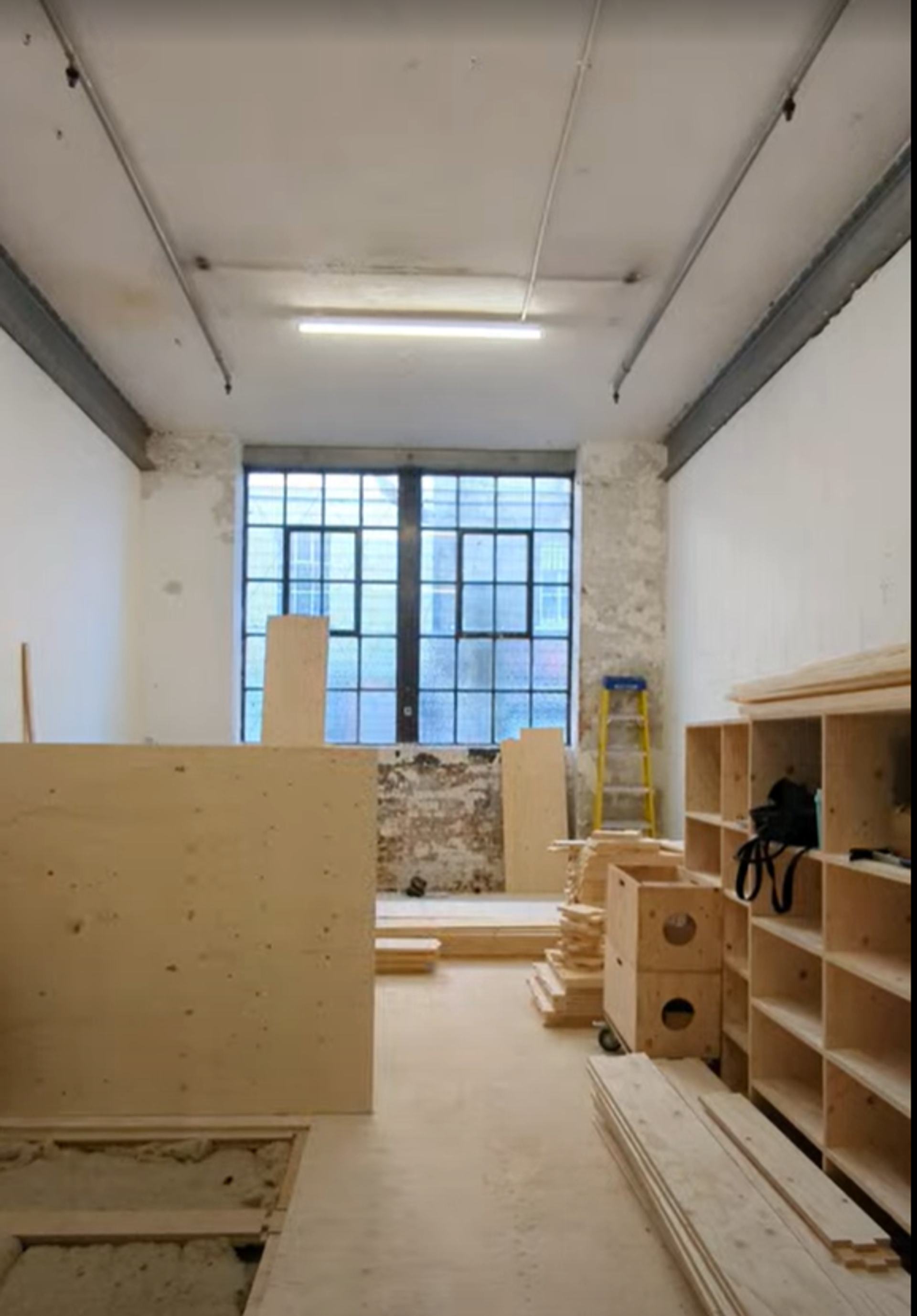 A workshop room with unfinished wooden shelves, boards, and plywood planks scattered around, a tall window at the far end, a yellow step ladder, and white walls with exposed, weathered brick by the window—perfect for sustainable timber construction projects.