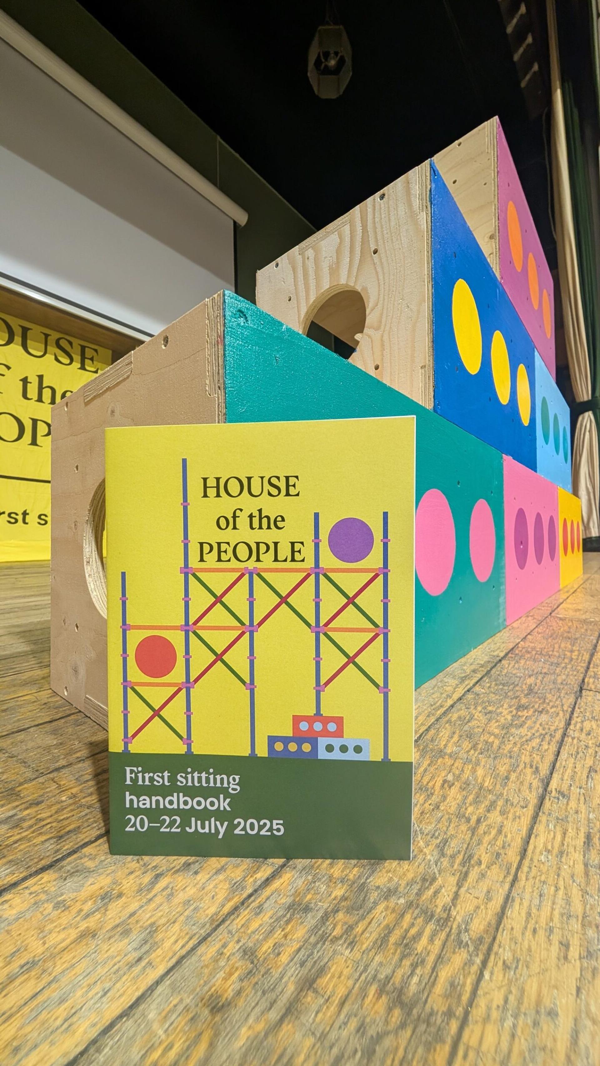 A colourful booklet titled “HOUSE of the PEOPLE: First sitting handbook 20–22 July 2025” stands on a wooden floor in front of a wooden structure painted with colourful circles, crafted from sustainably sourced materials.