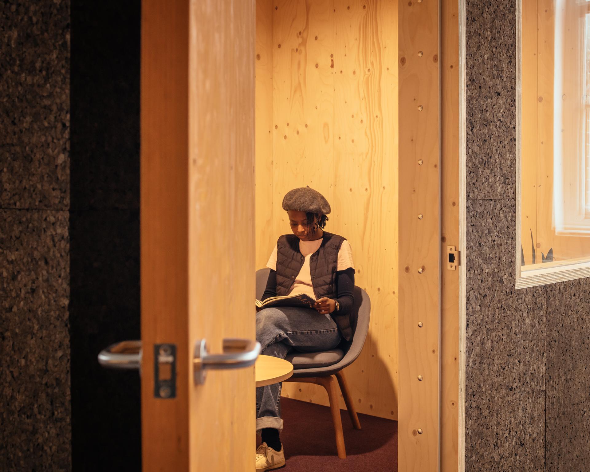 A person wearing a beret sits on a chair in a small birch timber-walled room, reading a book. The modular space is viewed through a partly open door, with a small round table in front of the person.