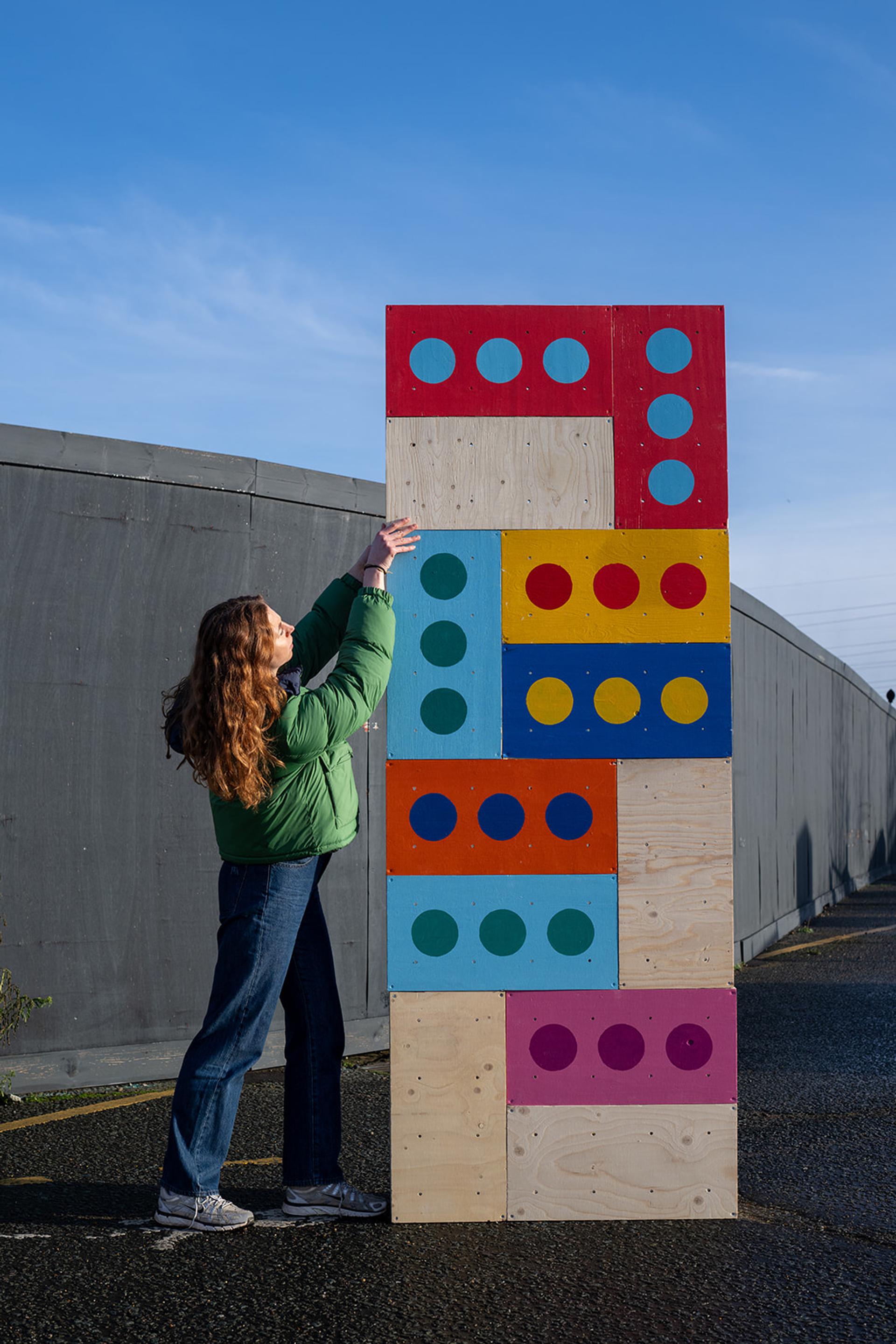 A woman in a green jacket and jeans reaches up to touch a tall, colourful wooden structure made of domino-like pieces, capturing a playful moment on the pavement outside what appears to be a Box Library under the blue sky.