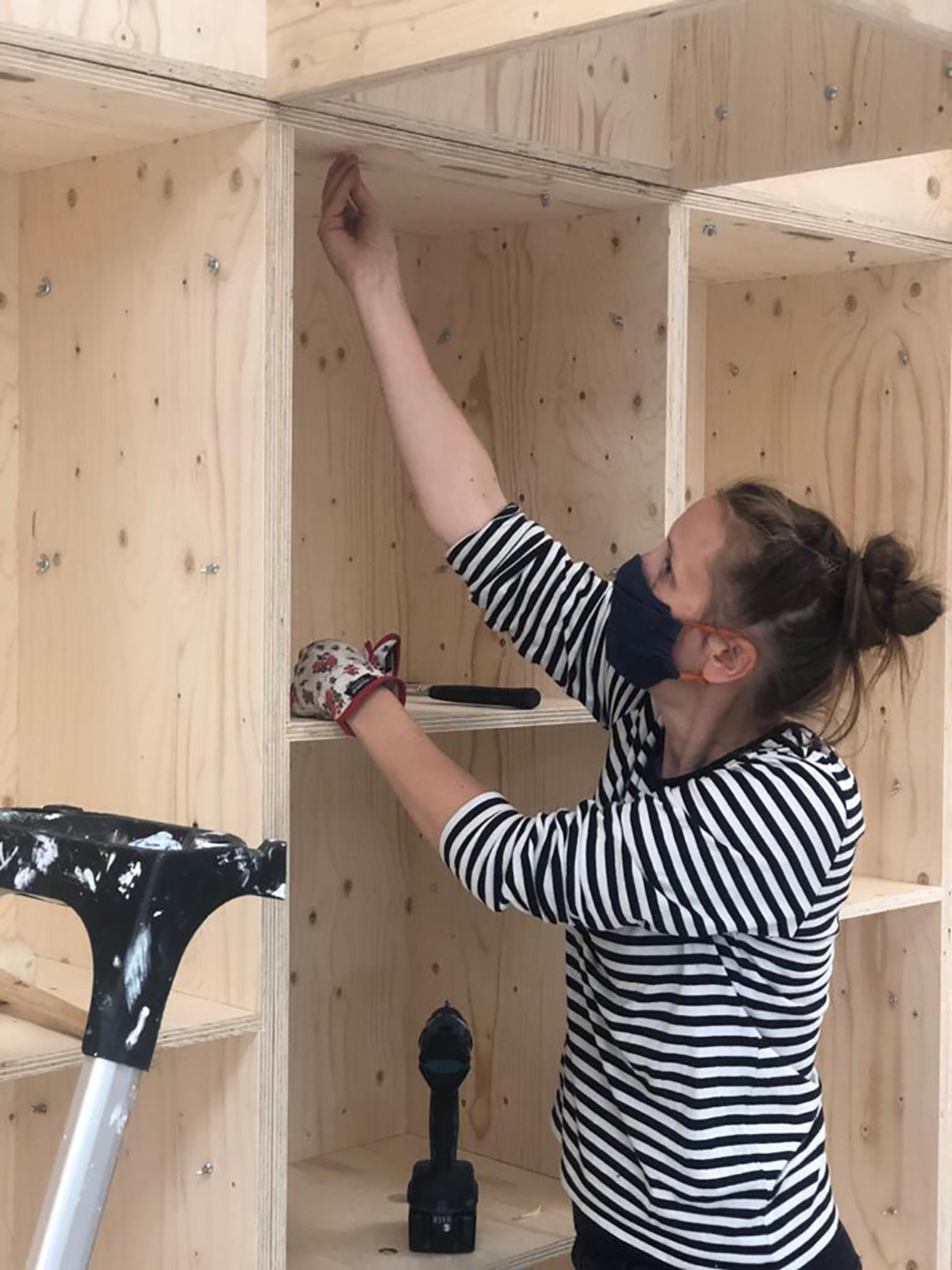 A woman wearing a striped shirt and a face mask installs or adjusts a screw in a modular wooden shelving unit. She is holding a tool, with a cordless drill and paint roller visible nearby, showcasing her skills in timber construction.