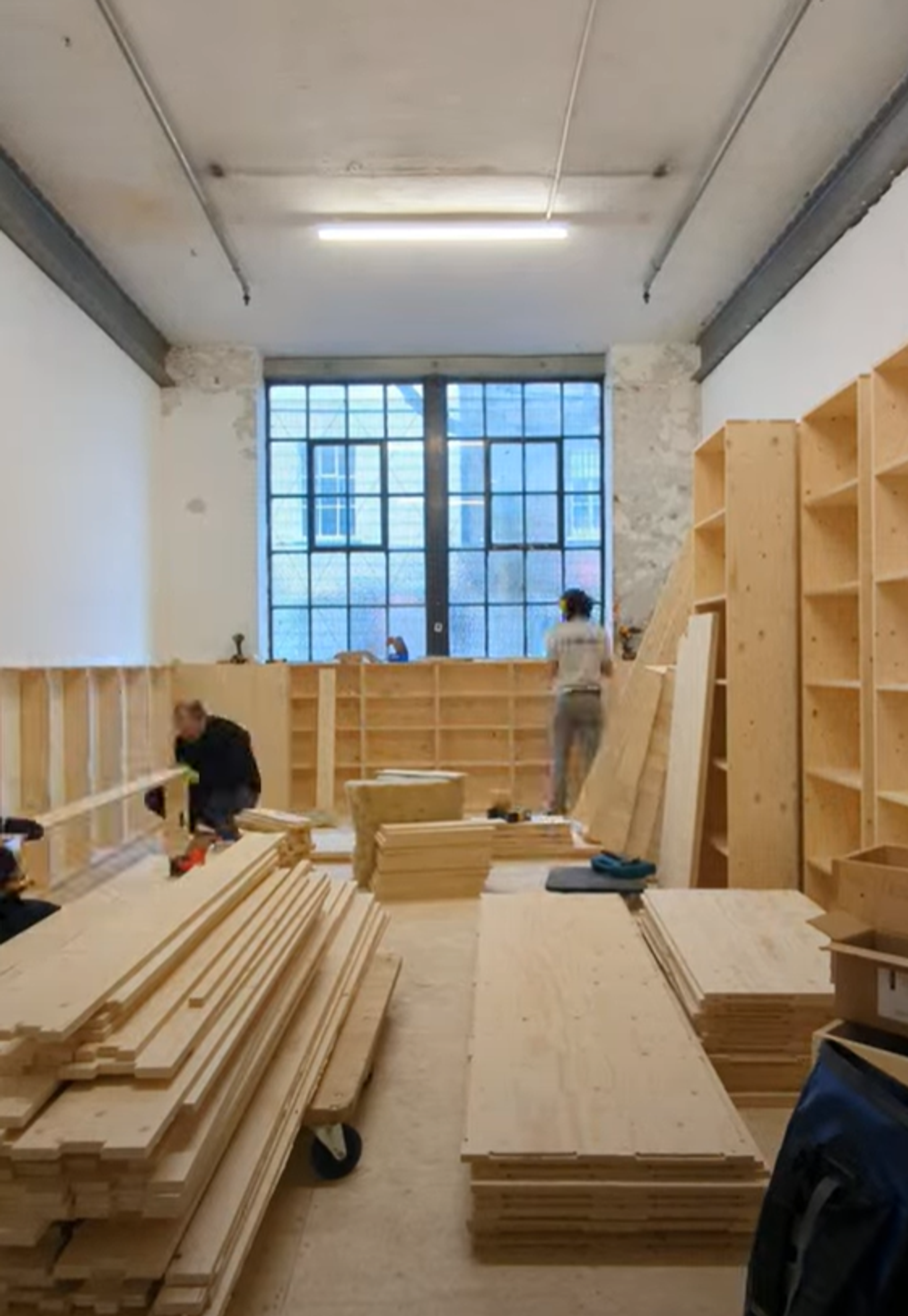 Two people assemble modular wooden shelves in a bright room with tall windows. Stacks of plywood planks and tools are scattered on the floor. Completed shelving lines the walls as light streams in from the large window at the back.