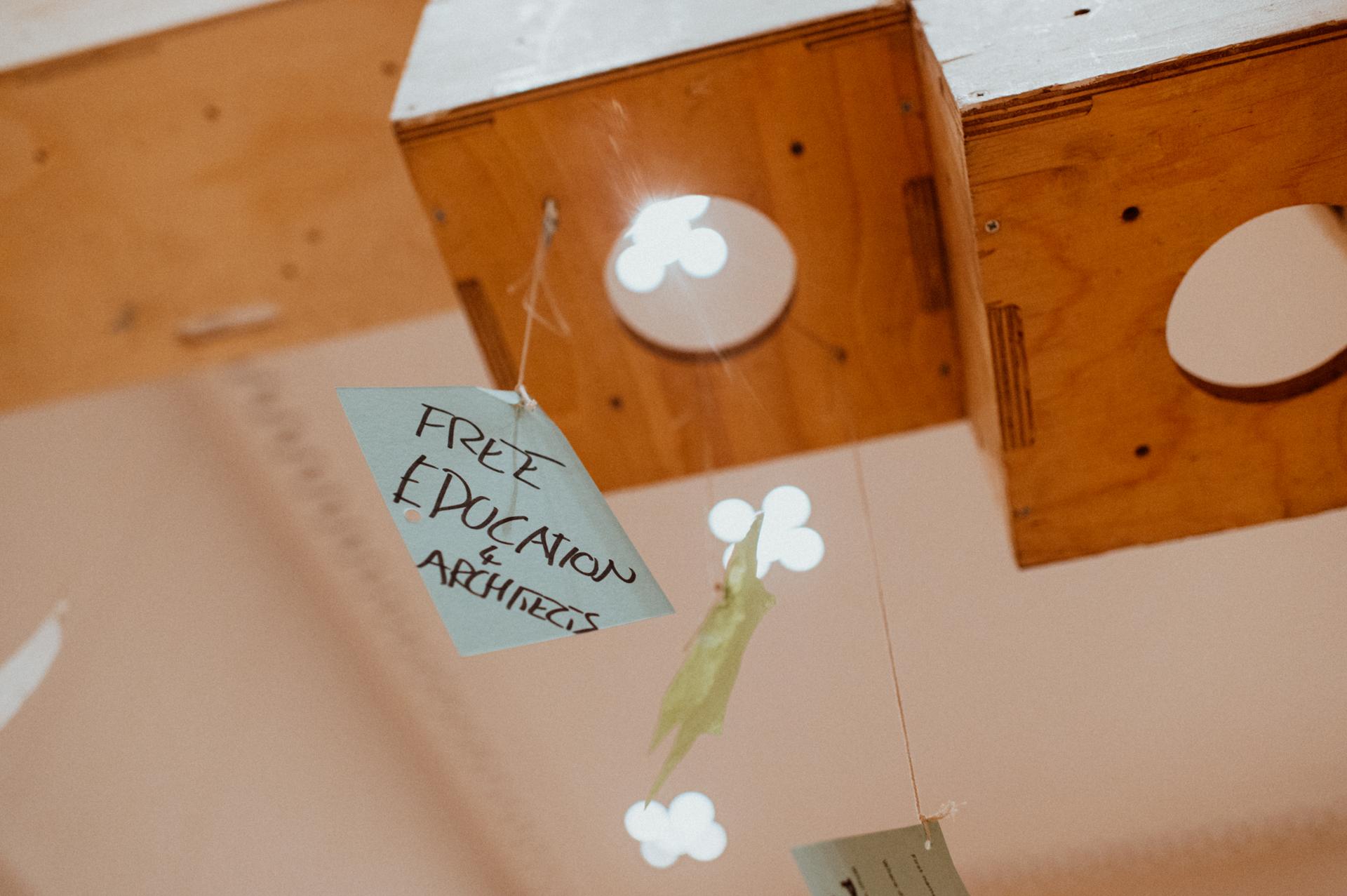 A close-up of a hanging paper sign reading FREE EDUCATION & ARCHIVES, attached to a string beneath a wooden community build project structure with flower-shaped cut-outs, highlighting creative youth architecture and design through plywood reuse.