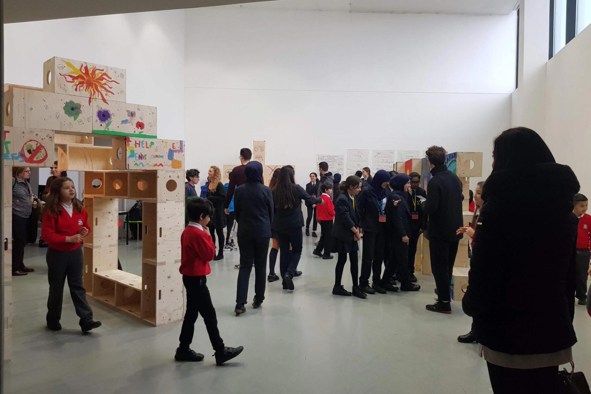 A group of children and adults gather in a bright gallery space with large, sustainably sourced art pieces and colourful drawings on the walls. Some pupils are in uniform, interacting and observing the artwork.