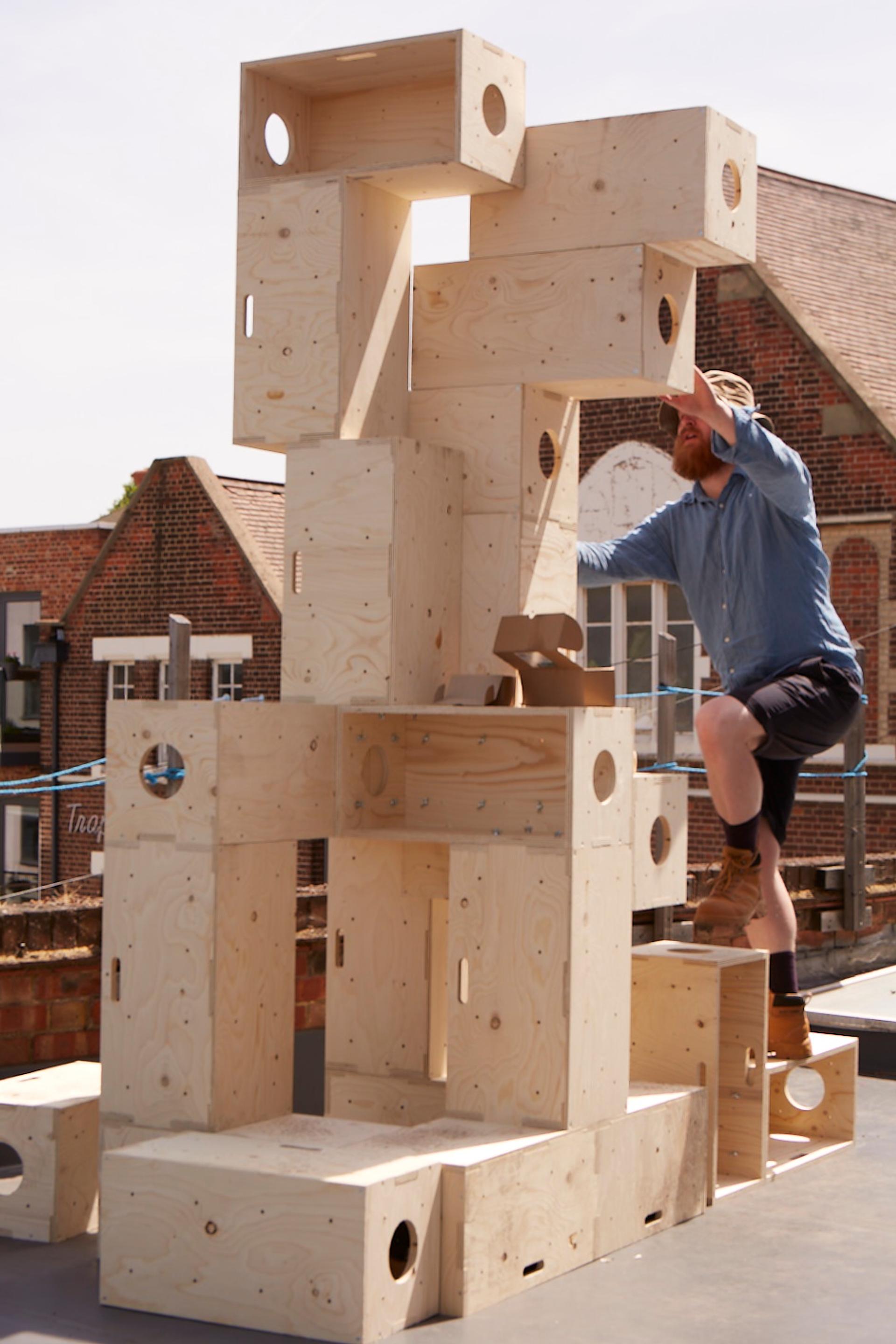 A man in a blue shirt and brown boots climbs a tall timber construction made of stacked spruce plywood boxes with circular cut-outs, outdoors on a sunny day with brick buildings in the background.