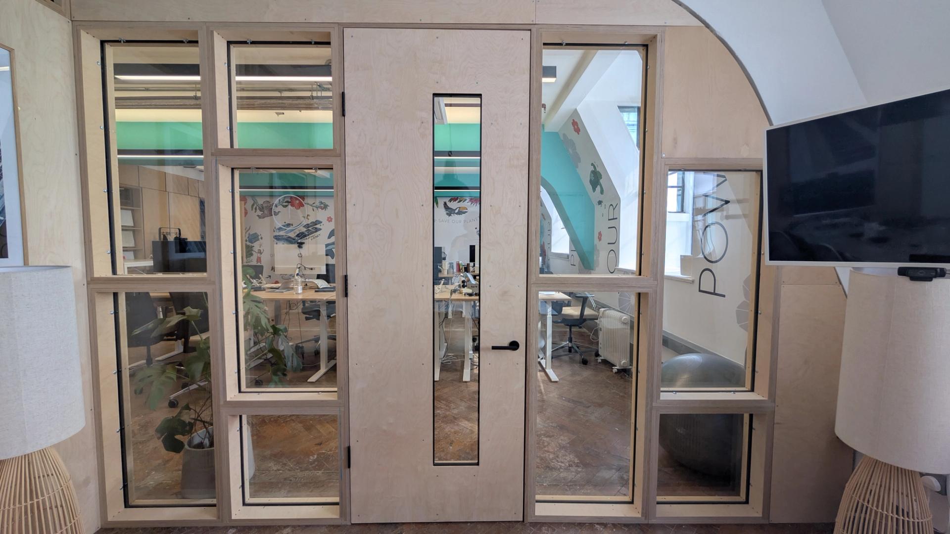A wooden-framed glass door, crafted from sustainable spruce, with side and top windows leads to a modern office space. Inside, desks, chairs, office supplies, and colourful wall art are visible. Two floor lamps and a TV are positioned outside the door.