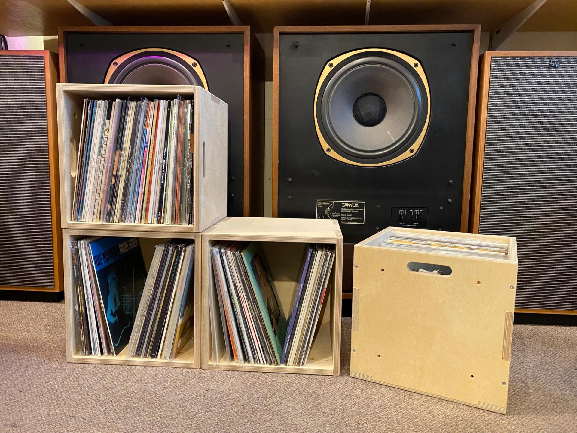 Four wooden crates filled with vinyl records are displayed on the floor in front of large black and brown speakers. The crates are arranged in a staggered formation on a carpeted surface.