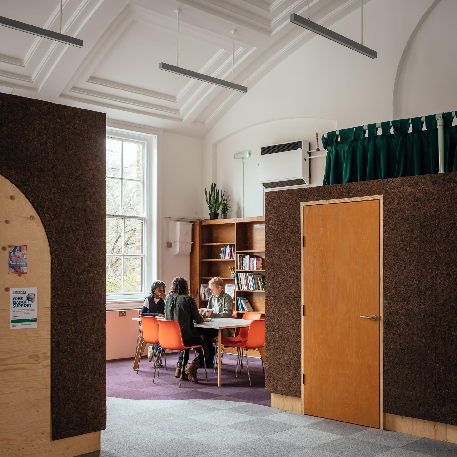 Three people sit together at a table in a modern library or study area with bookshelves, orange chairs, birch accents, a large window, and overhead lighting. The space features sustainable timber construction, high ceilings, and wooden doors.
