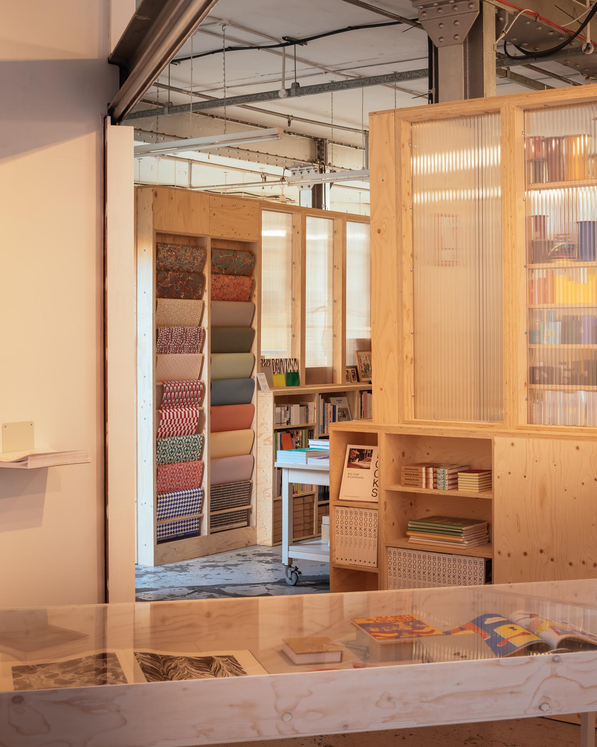 A cosy, modern shop interior with birch plywood shelves, vibrant patterned rolls of paper, books, and a glass display case. Sunlight softly illuminates the modular space, creating a warm and inviting atmosphere.