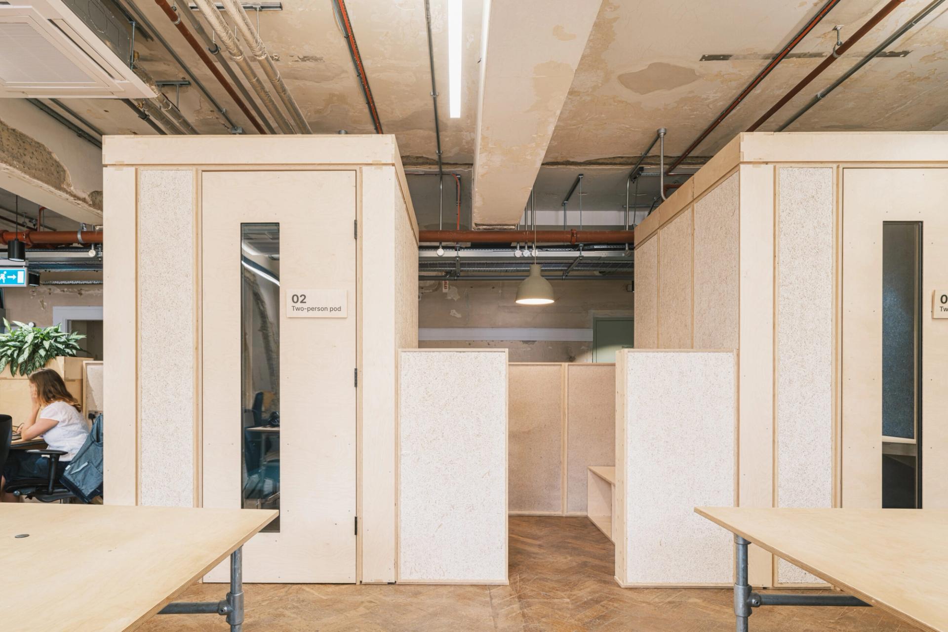 A modern office space with sustainable wooden meeting pods, exposed ceilings, and hanging lights. A person is working at a desk on the left, whilst the area on the right features empty desks and birch seating.