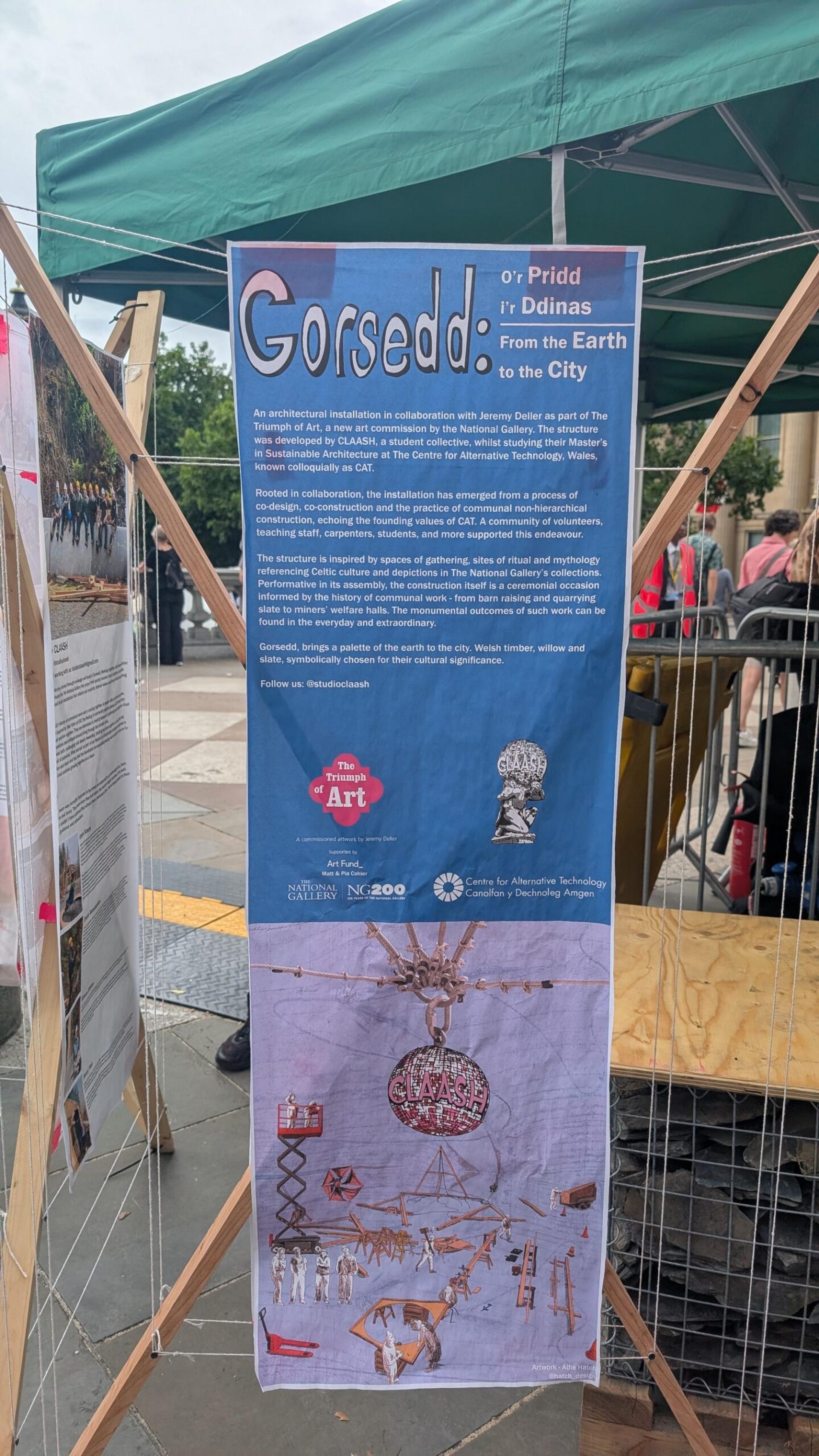 A tall blue poster titled Gorsedd: From the Earth to the City is displayed on a wooden frame at an outdoor art exhibition. The 2025 poster features English and Welsh text, logos, and artwork of sculptural objects on the lower half.