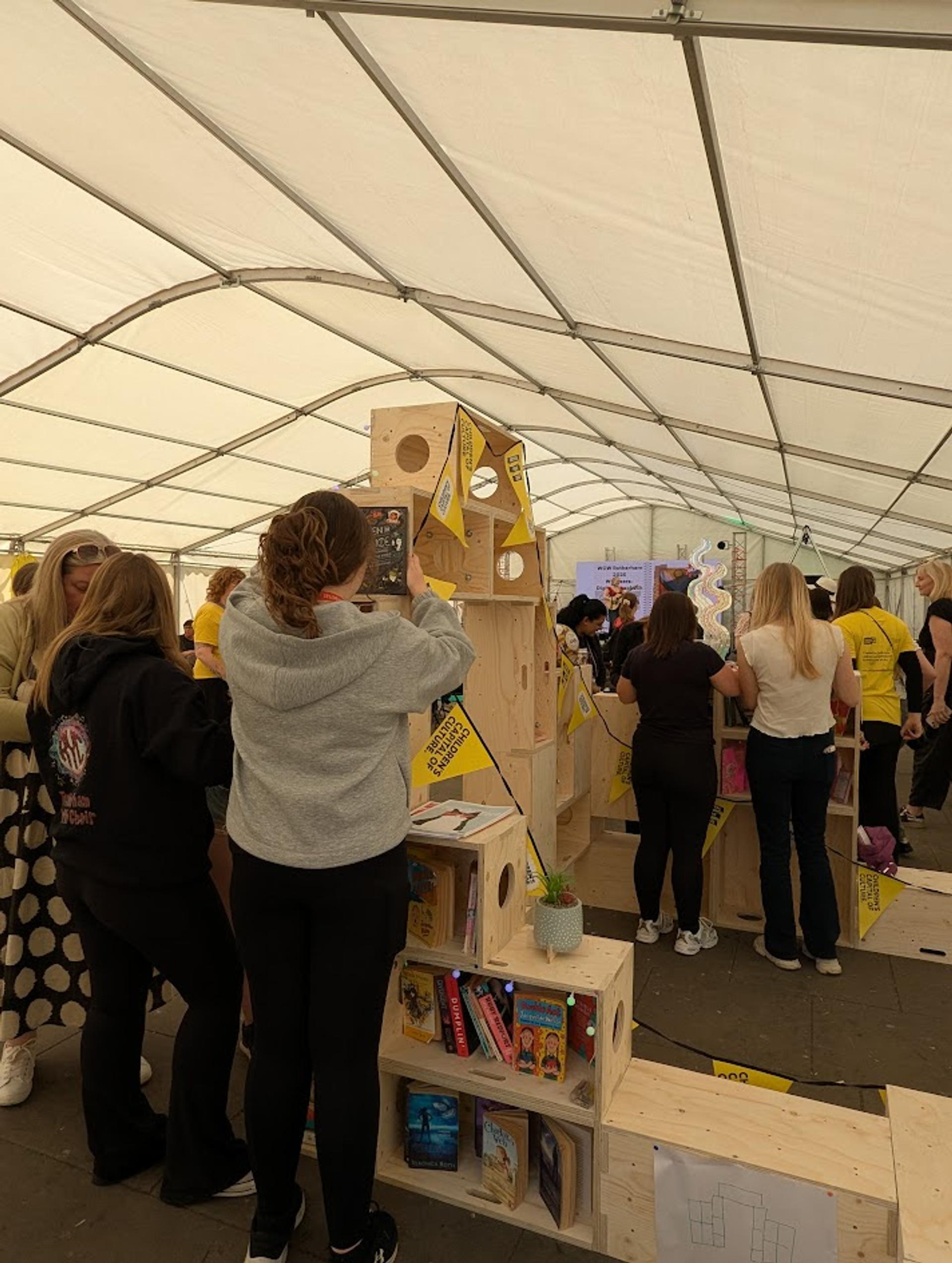People gather under a large white marquee, interacting with sustainable wooden structures filled with books, decorations, and yellow flags. Some individuals are taking photos while others are socialising in groups.