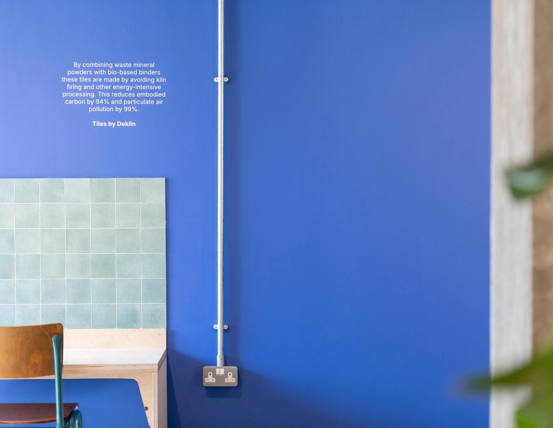 A turquoise-tiled table with a wooden chair sits against a vibrant blue wall in Manchester. A metal conduit runs vertically, and white text about Sustainable Ventures and sustainable business tile production is displayed above the table.