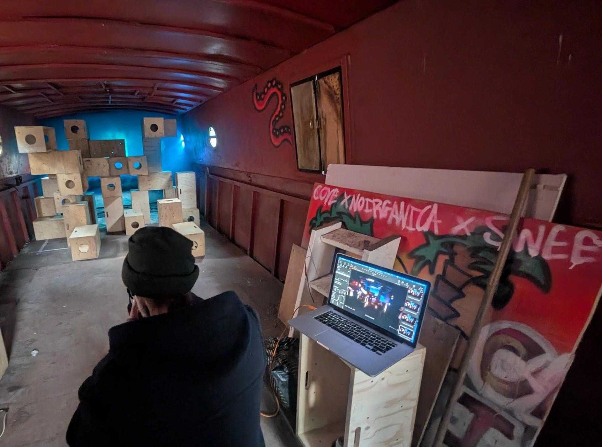 A behind-the-scenes wide shot of the industrial space. In the foreground, photographer Vincent looks toward the modular wooden stack in the distance. To the right, a laptop sits on a wooden crate displaying photo editing software, next to a large piece of plywood leaning against the wall covered in red and pink graffiti. The background features U-Build boxes illuminated by a cool blue light.