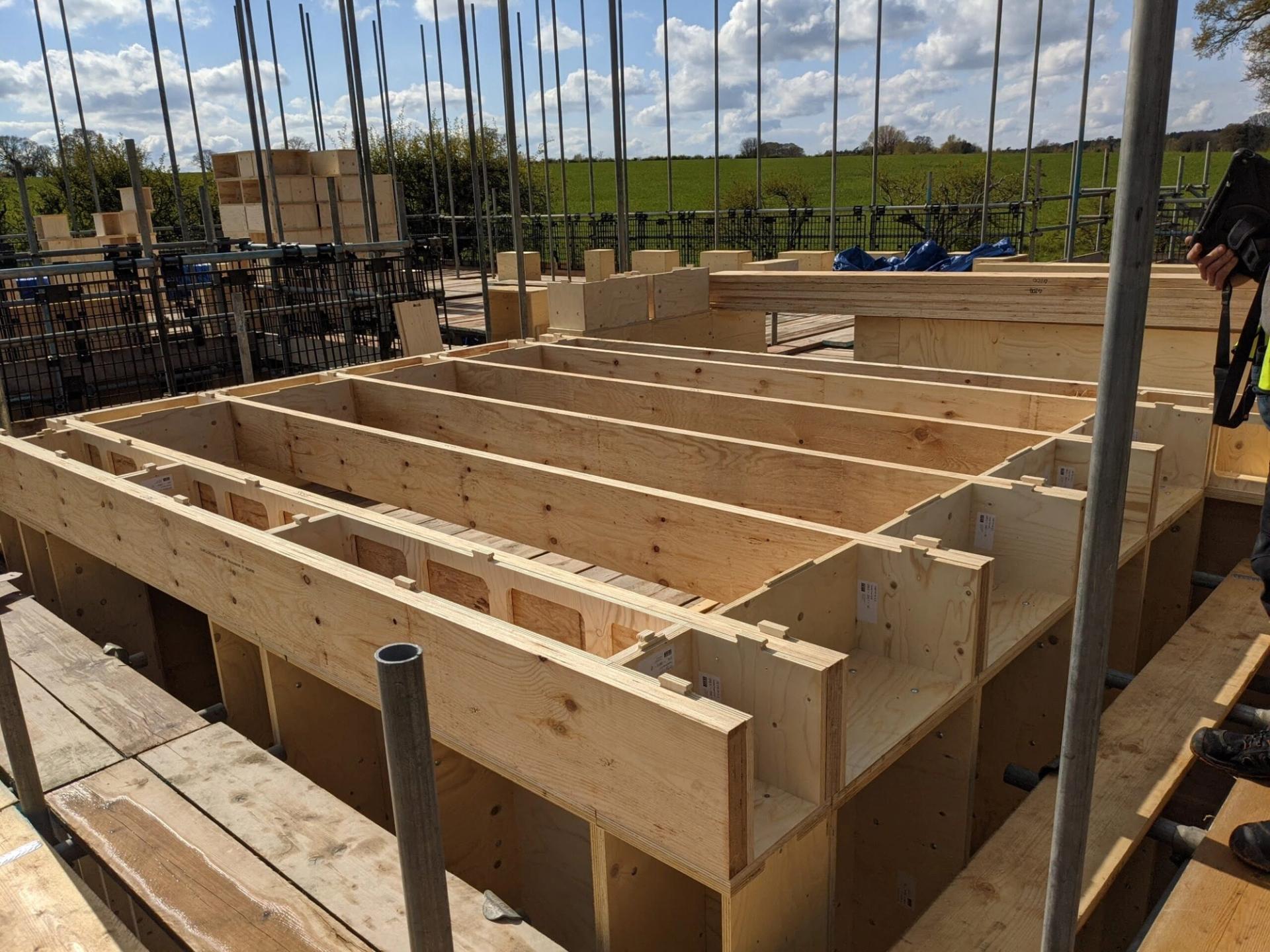 A construction site with spruce timber framing for a building foundation, surrounded by scaffolding and metal poles, in an outdoor rural area with green fields and a blue sky with clouds in the background.