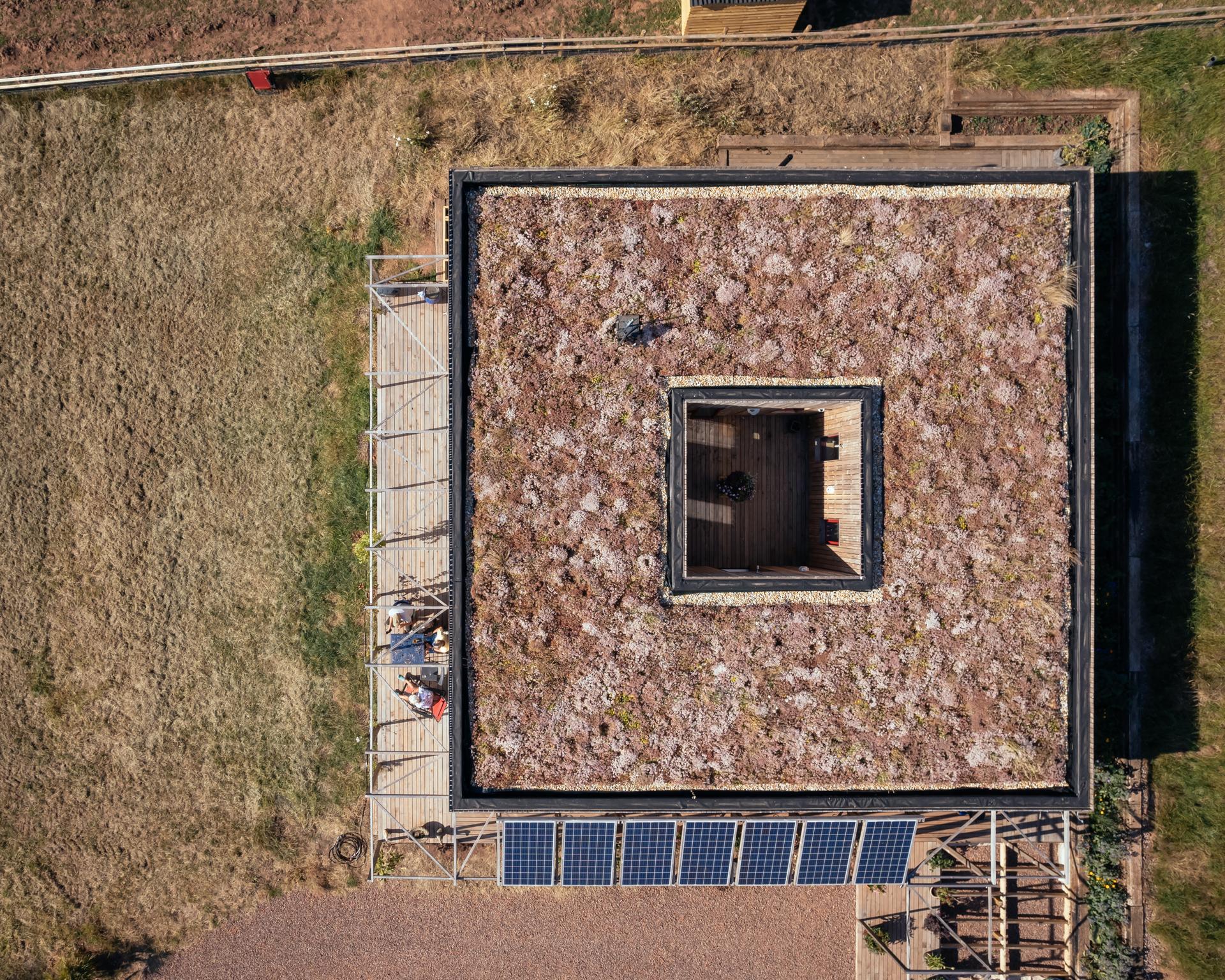 Aerial view of a modular square building with a green roof and central open courtyard. Solar panels line one side, whilst people sit on a terrace surrounded by dry grass, highlighting the use of sustainable timber construction.