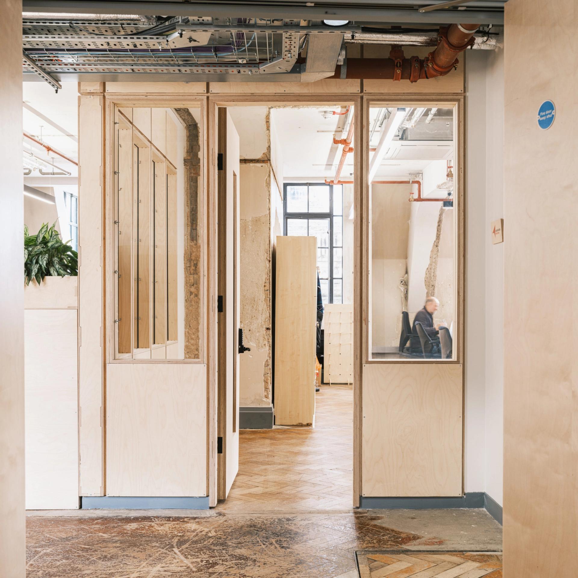 A modern office space with light spruce wooden doors and glass panels, exposed pipes on the ceiling, and a person sitting at a desk in the background near large windows showcase an elegant touch of modular design.