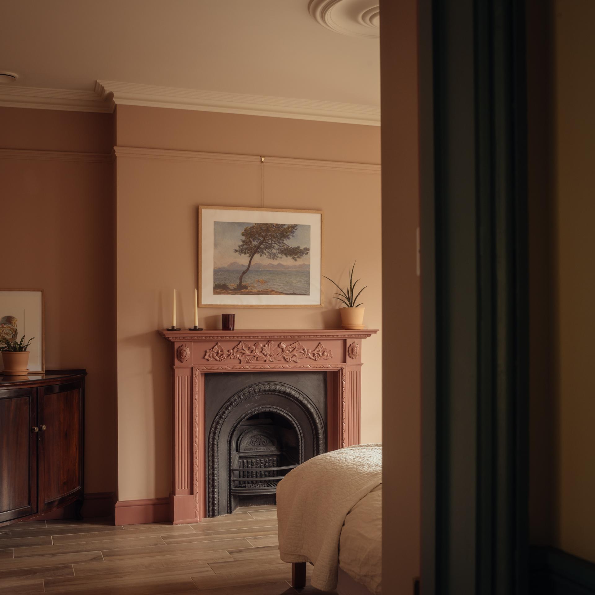 An ornate timber fireplace mantel topped by a framed landscape painting, flanked by candles and a small potted plant.