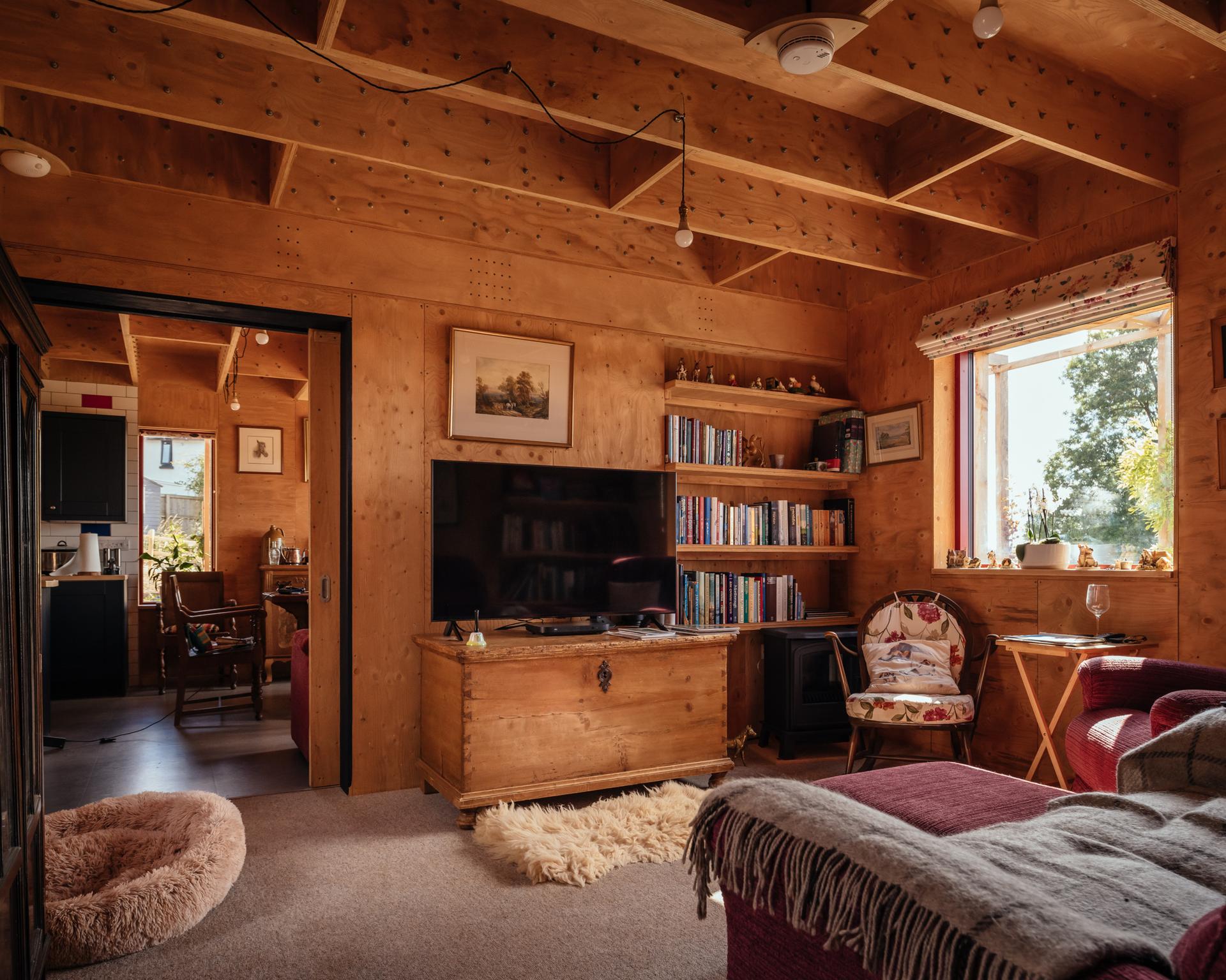 Cosy spruce-panelled living room with exposed beams, a TV on a chest, bookshelves, armchairs, a pet bed, and sunlight streaming through a window. An open doorway leads to the kitchen and dining area.