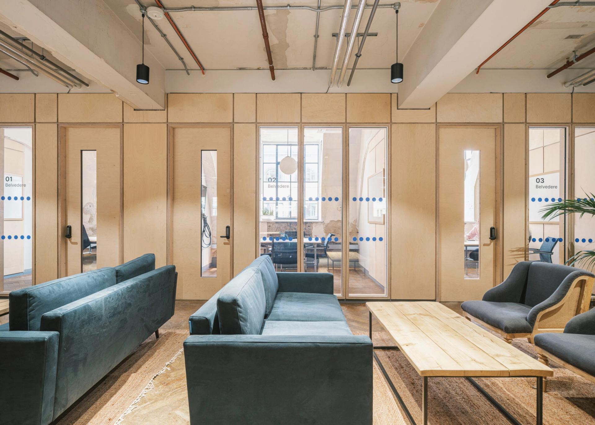 Modern office lounge with blue sofas, a birch coffee table, and glass-walled meeting rooms in the background. The space features exposed ceilings, light wood walls, and natural light from large windows.