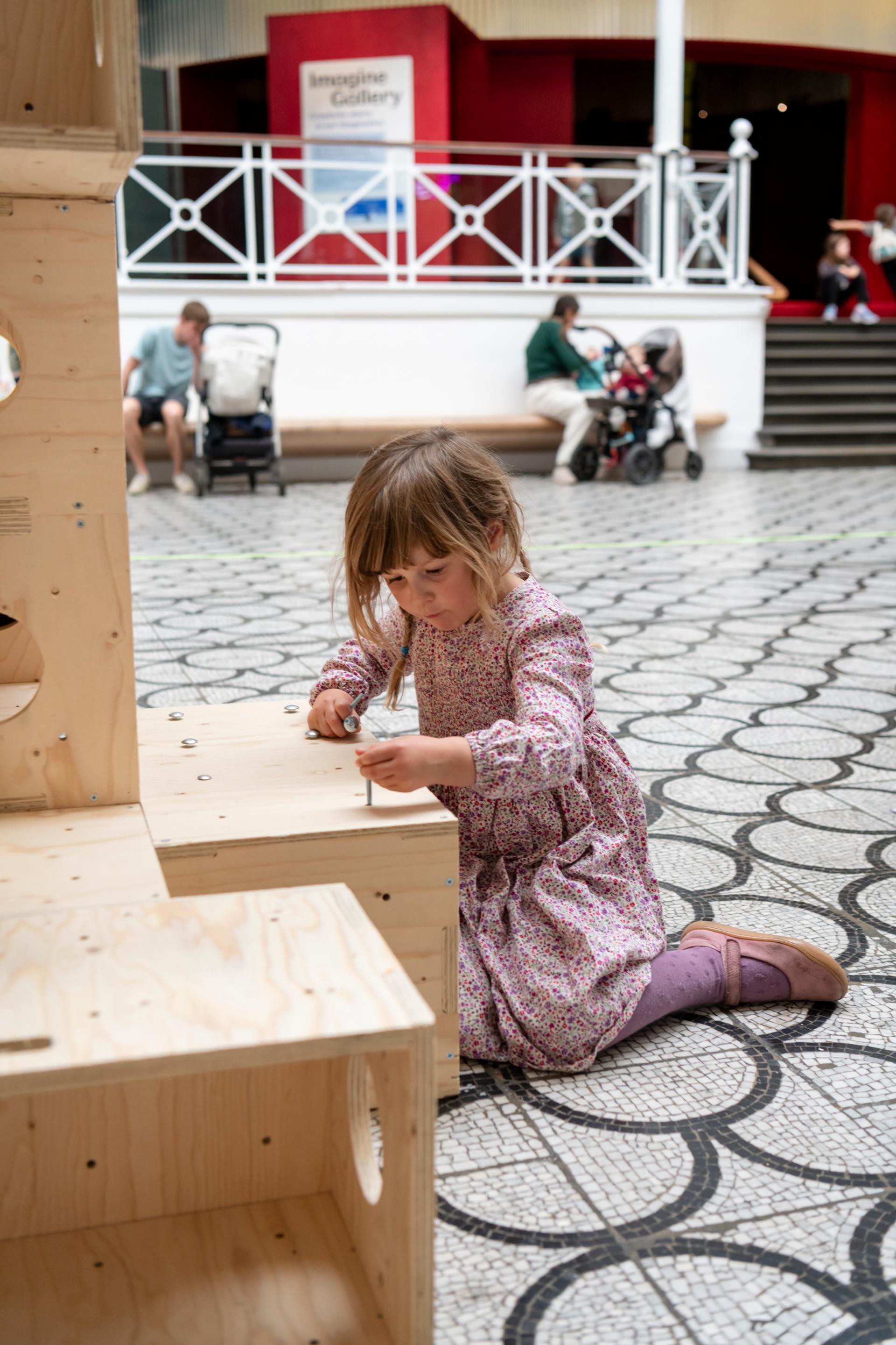 A young girl in a floral dress kneels on a patterned floor, using U-Build tools to assemble a wooden structure. People sit in the background near a white-railed area with a sign reading Imagine Gallery.