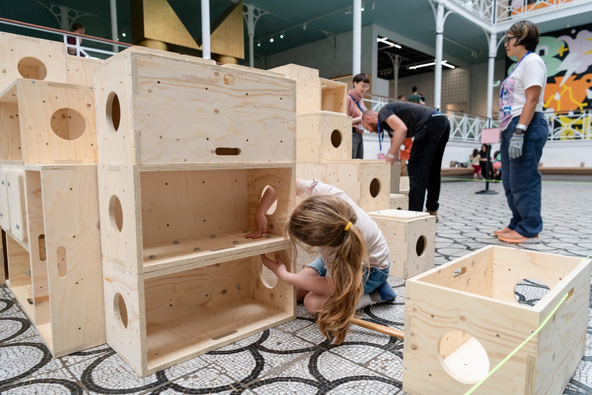 A young girl with long hair builds with large U-Build wooden boxes in an indoor space. Other people work nearby on a tiled floor, surrounded by more wooden structures and colourful wall art.