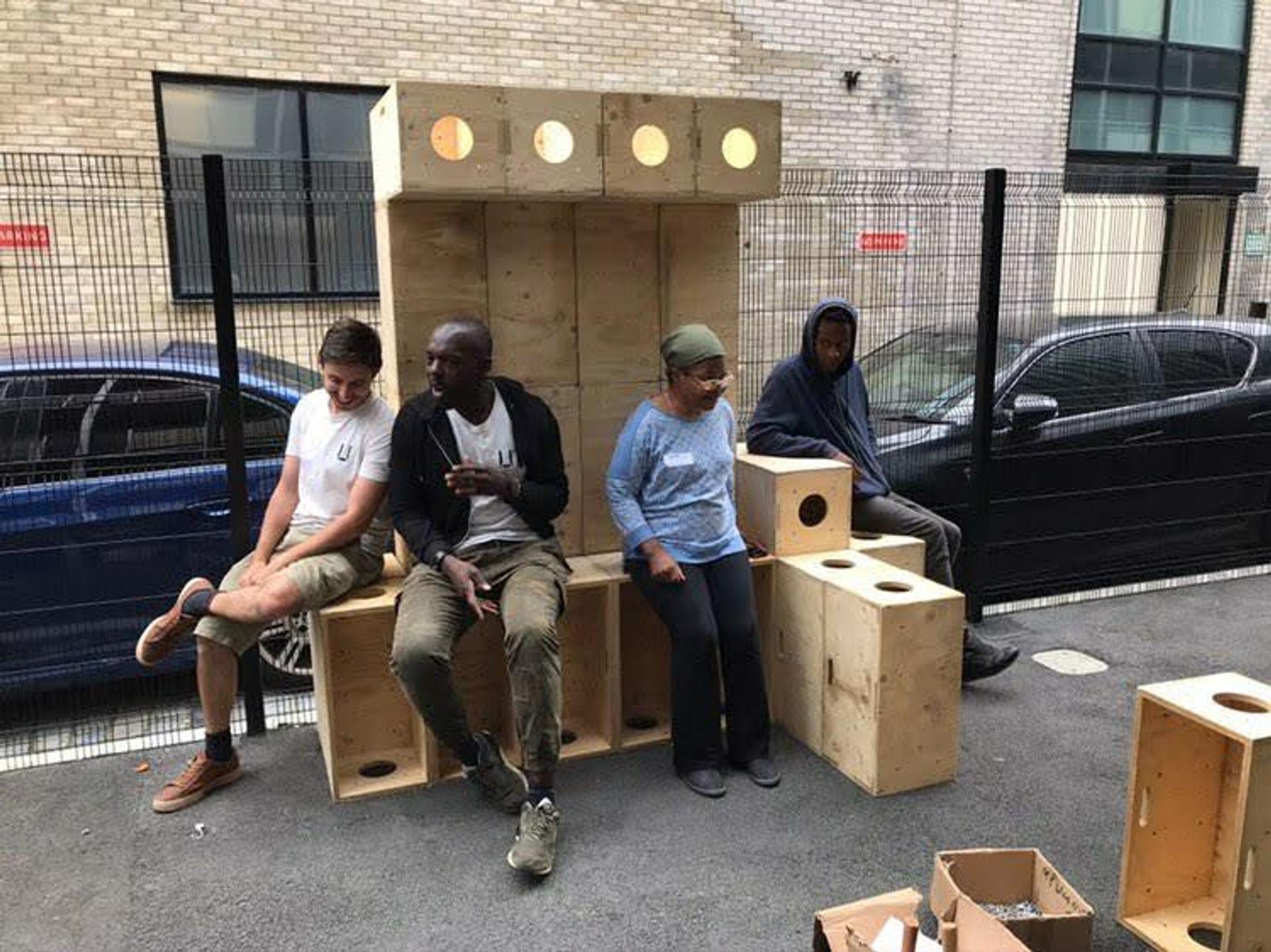 Four people sit on and beside large, sustainably sourced wooden box structures outdoors near a street with parked cars and a brick building. Some boxes feature circular cut-outs, with upcycled cardboard and other materials scattered on the ground.