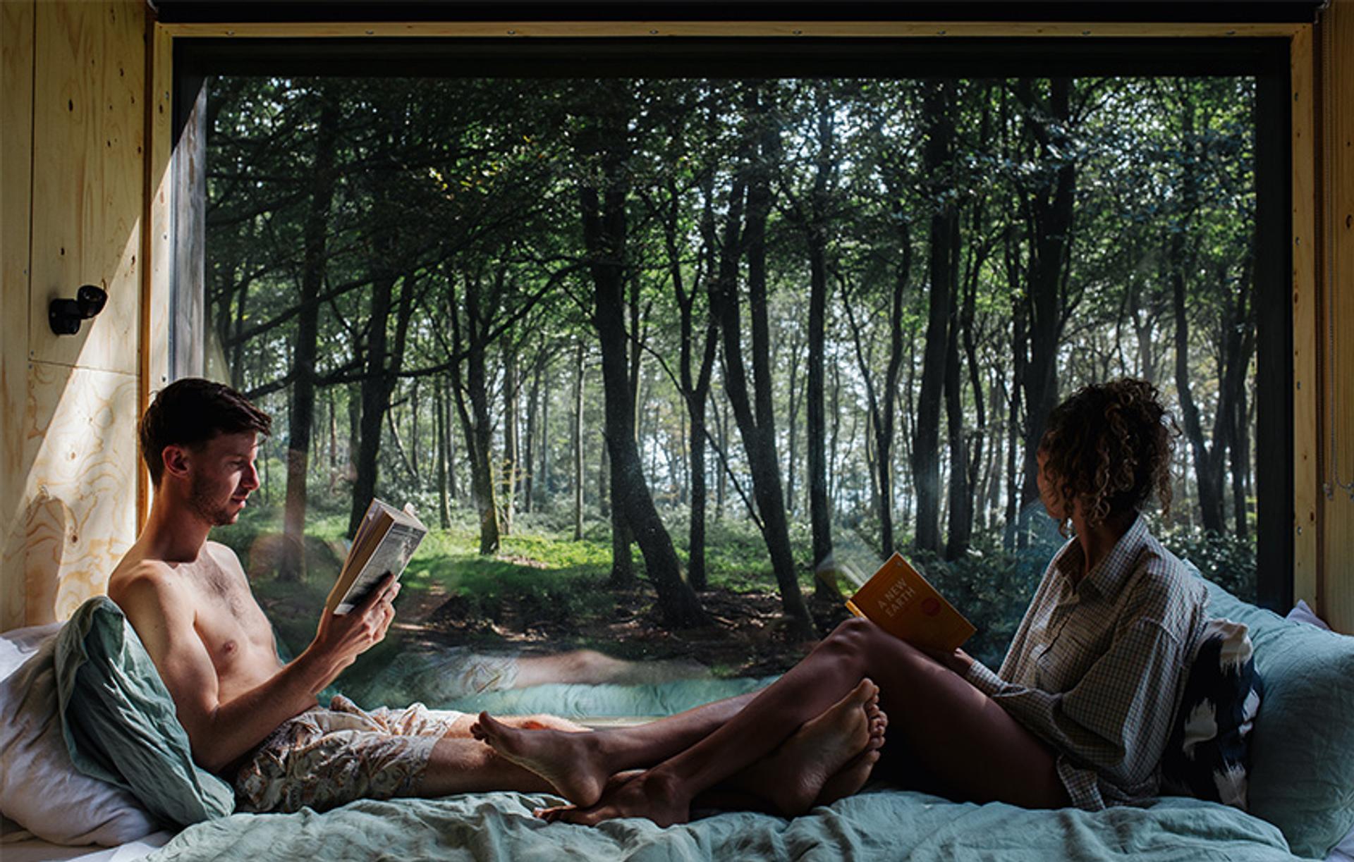 A man and woman sit on a bed in a cosy wooden cabin, reading books. Surrounded by upcycled decor, they enjoy the warmth as sunlight pours through a large window, revealing a lush forest just outside.