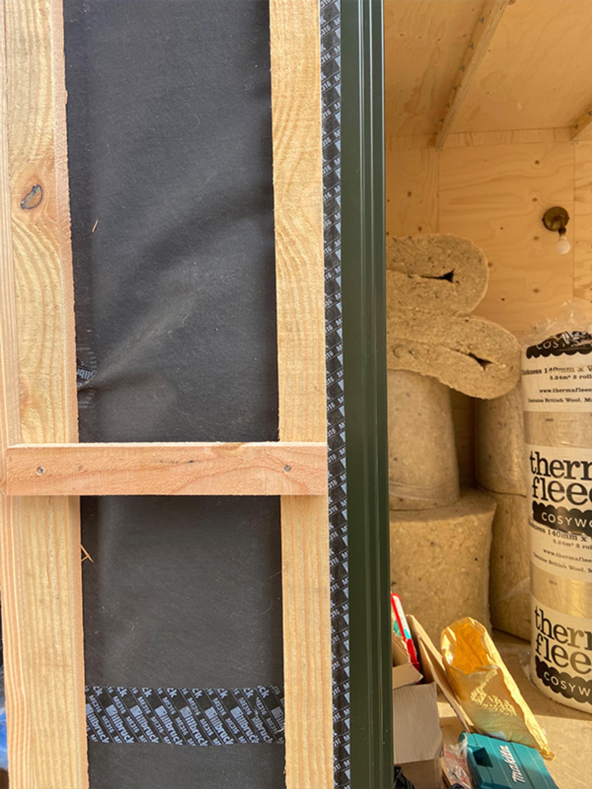 A close-up of a partially constructed wall with spruce wooden framing, black insulation, and tape. In the background, rolls of Thermafleece insulation and various construction materials are visible inside a wooden shed.