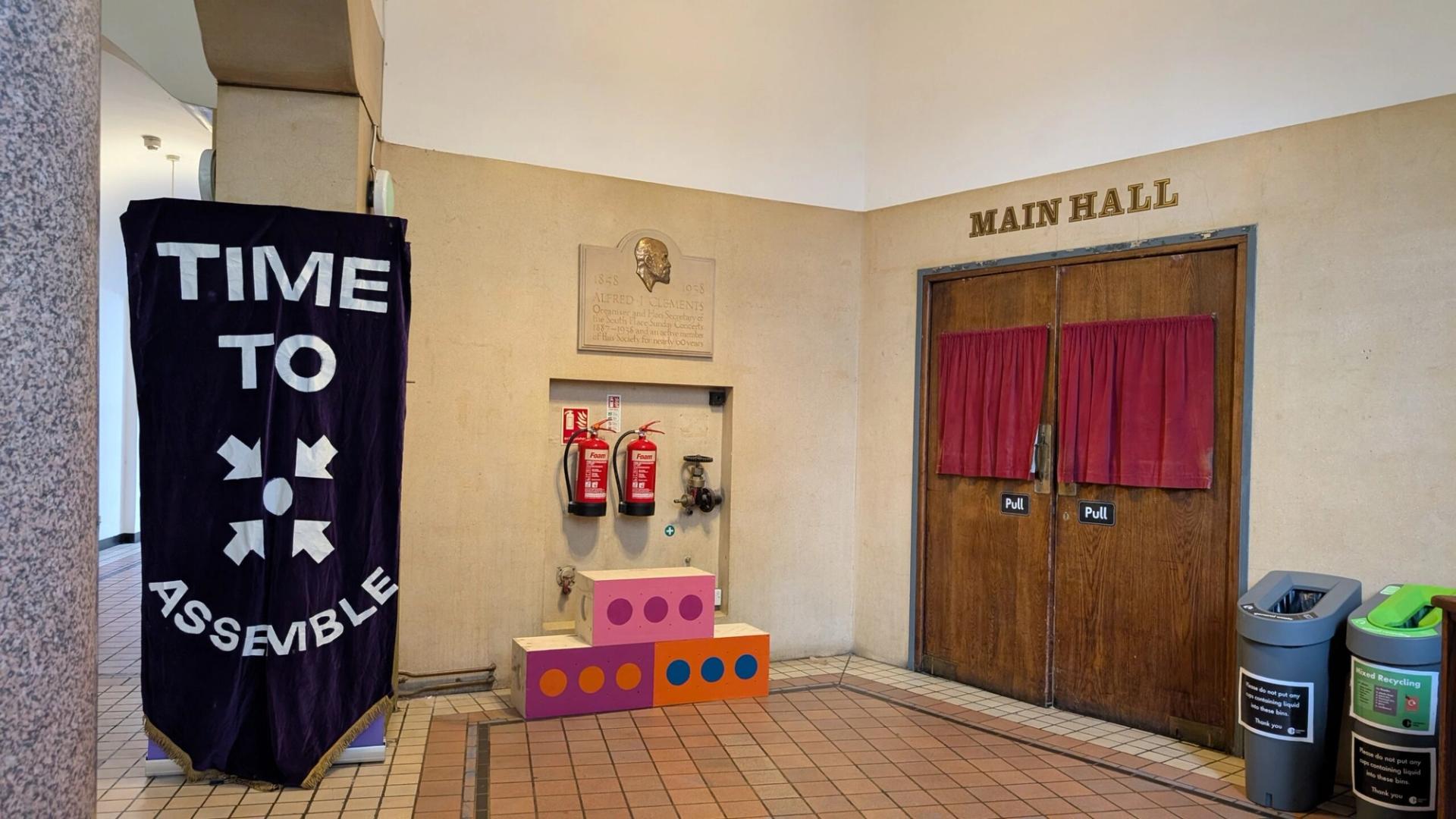 A banner reading TIME TO ASSEMBLE hangs near a wall with fire extinguishers, colourful upcycled blocks, and double doors labelled MAIN HALL. Two recycling bins are on the right side of the doors.