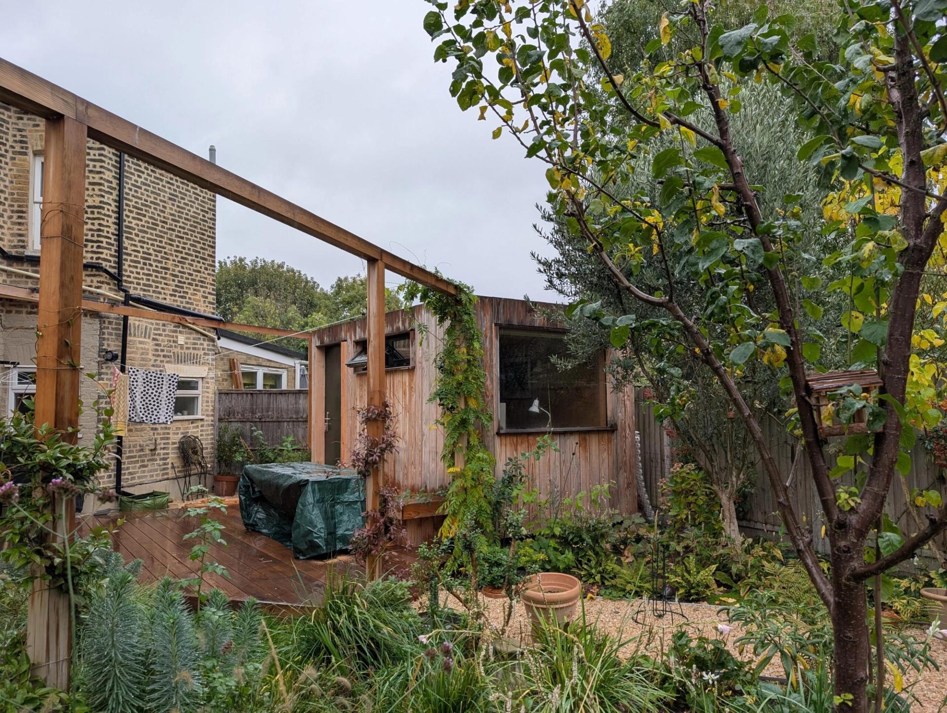 A lush back garden with various green plants and trees surrounds a small wooden U-Build shed. A covered object sits on a wooden decking next to a brick house on a cloudy day.