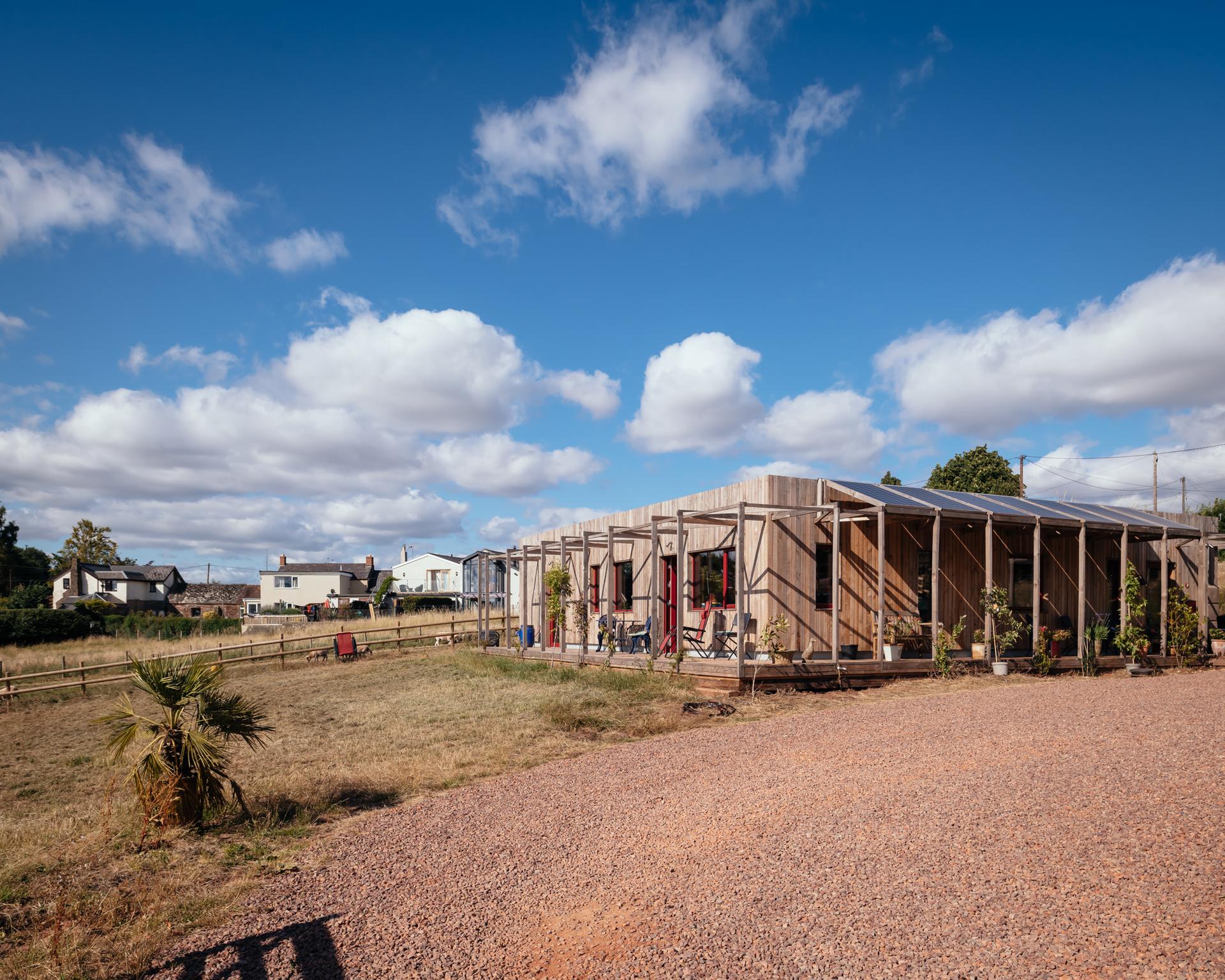 A modern, single-storey spruce building with large windows sits on a gravel car park, surrounded by dry grass and a few small plants under a bright blue sky with scattered clouds. Houses and trees are visible in the background.