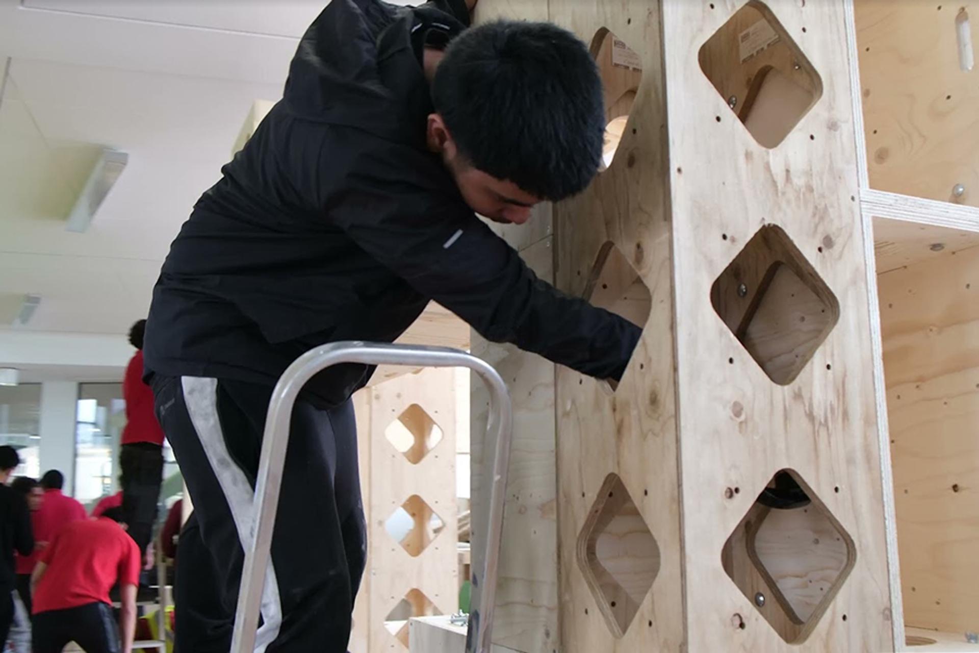 A person stands on a stepladder, working with their hands inside a large birch ply structure with diamond-shaped cut-outs; other people are visible in the background.