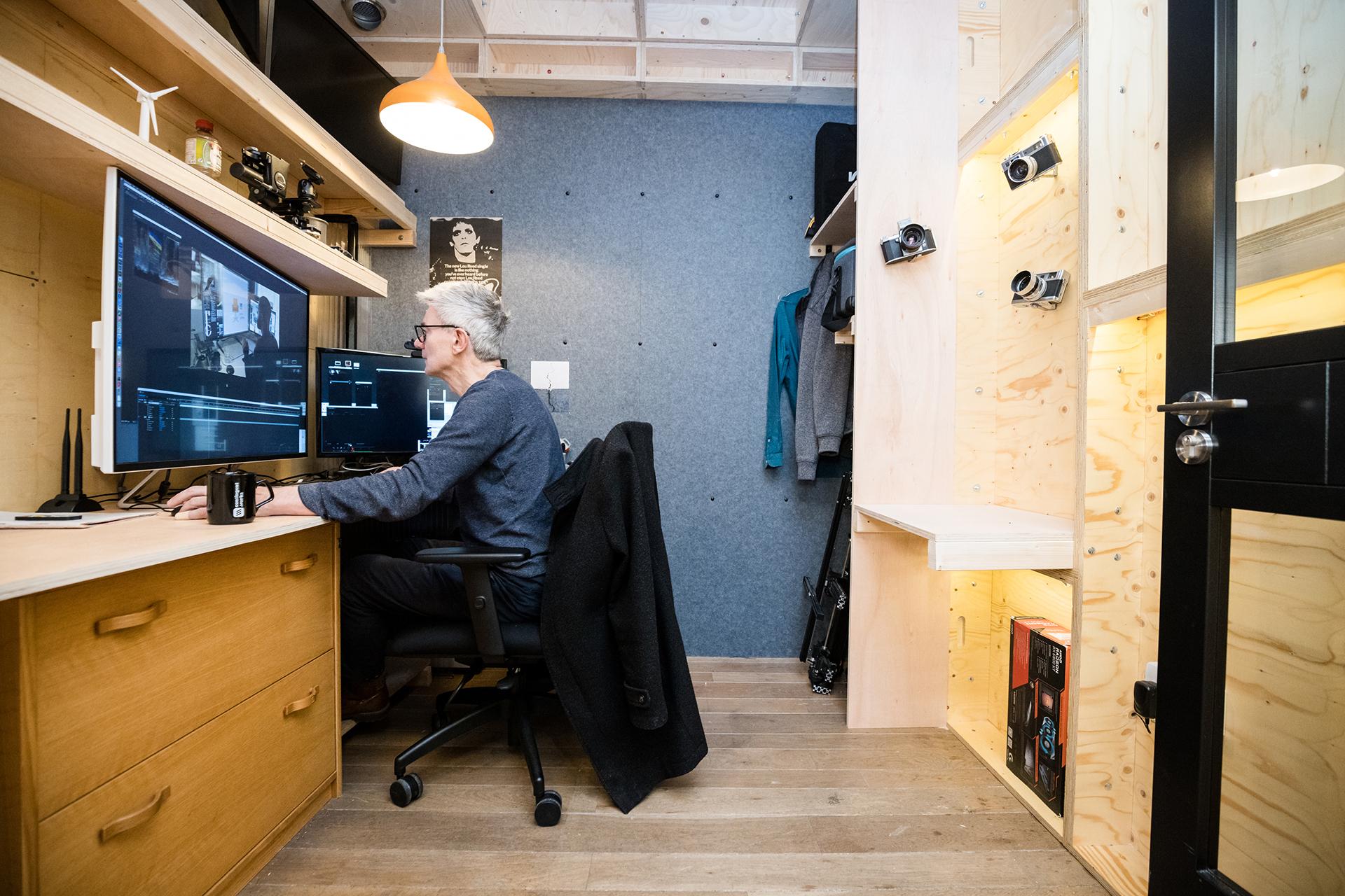 A person with grey hair and glasses edits video at a dual-monitor desk in a modern, organised workspace with birch plywood shelves, cameras on display, and a coat hanging nearby.