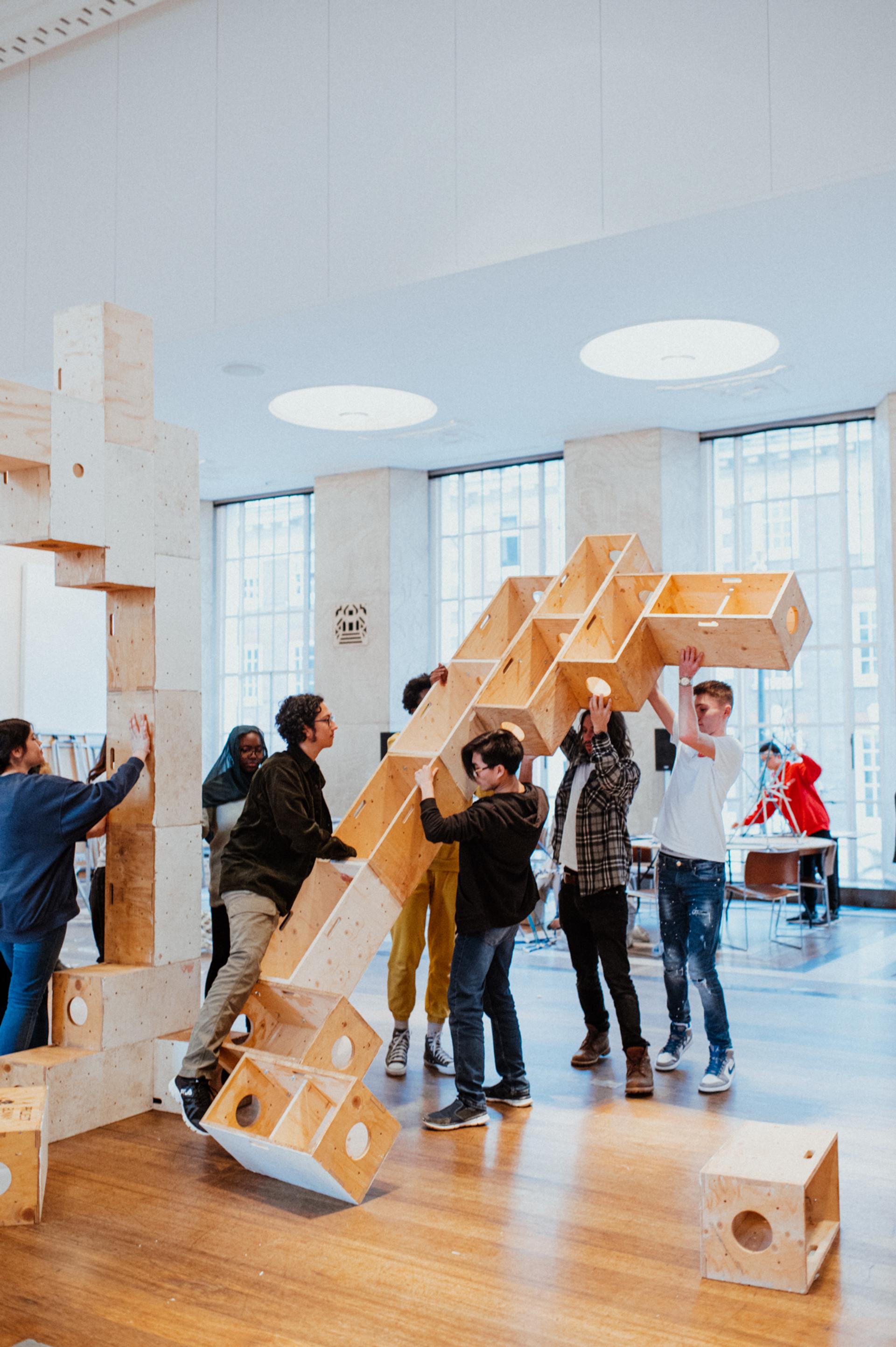 A group of young people work together to lift and assemble large plywood blocks inside a bright room with tall windows and wooden floors.
