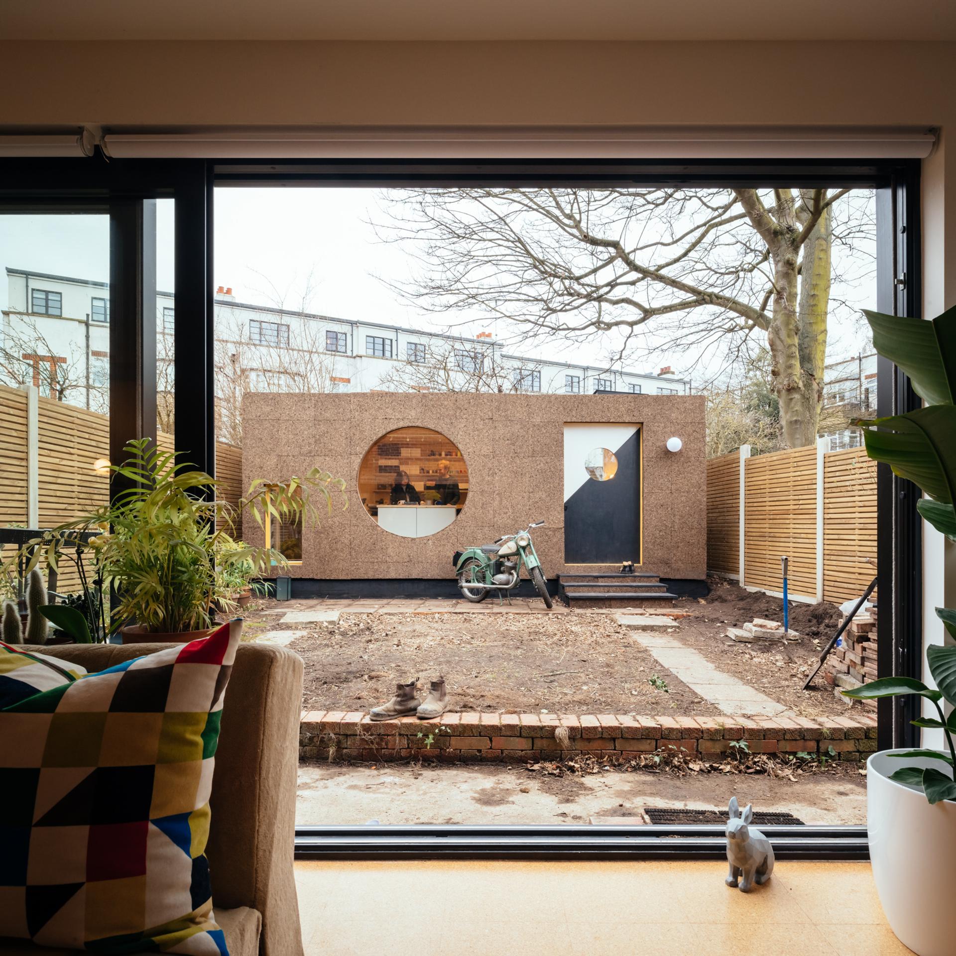 A view through a large glass door shows a modern garden office with a round window and black door. Spruce plywood accents frame the scene, while a scooter and gardening tools rest outside, and potted plants decorate the cosy interior.