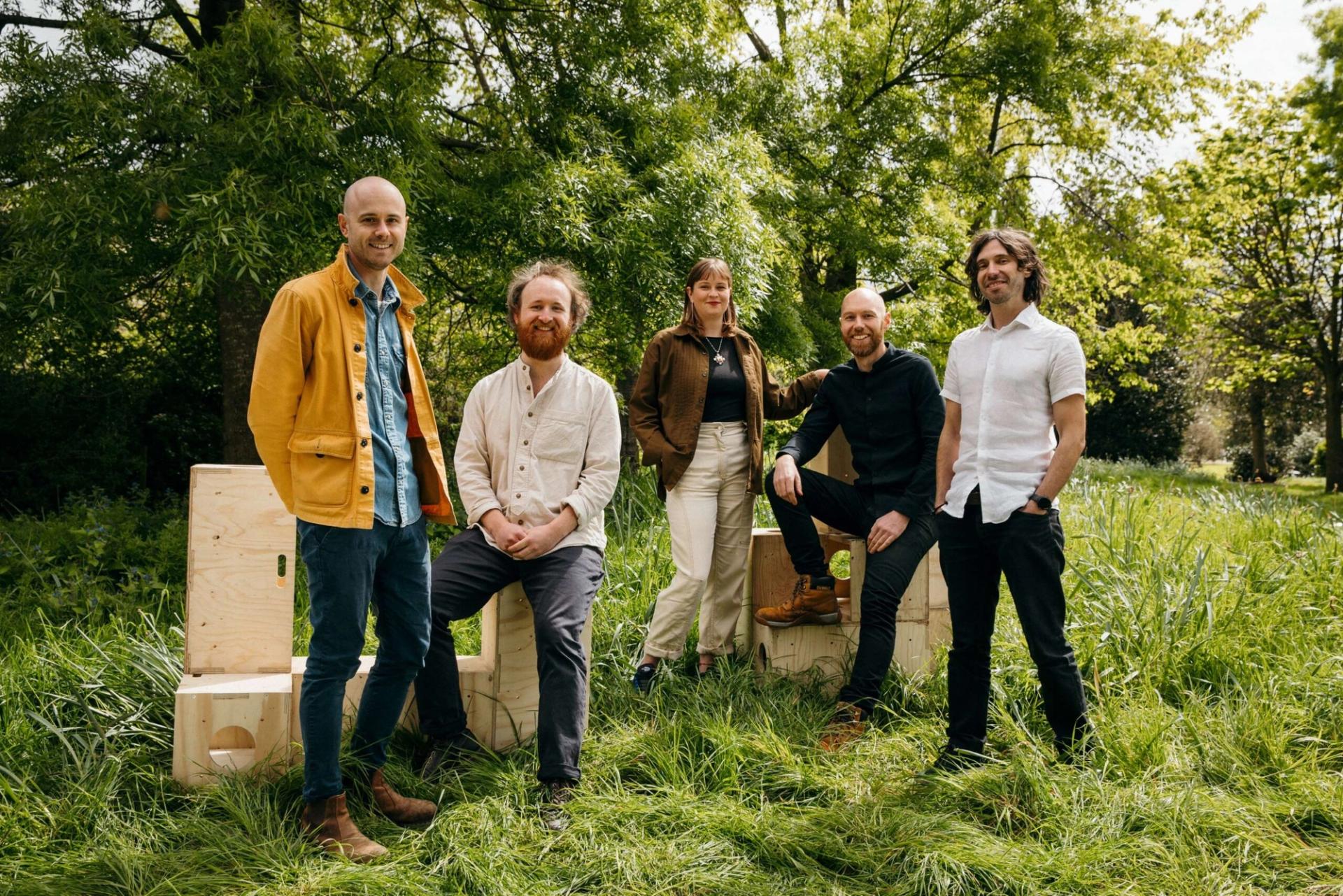 Five adults stand and sit on modular plywood boxes outdoors in a grassy, green park, surrounded by trees. They are casually dressed and smiling, posing together in natural sunlight.