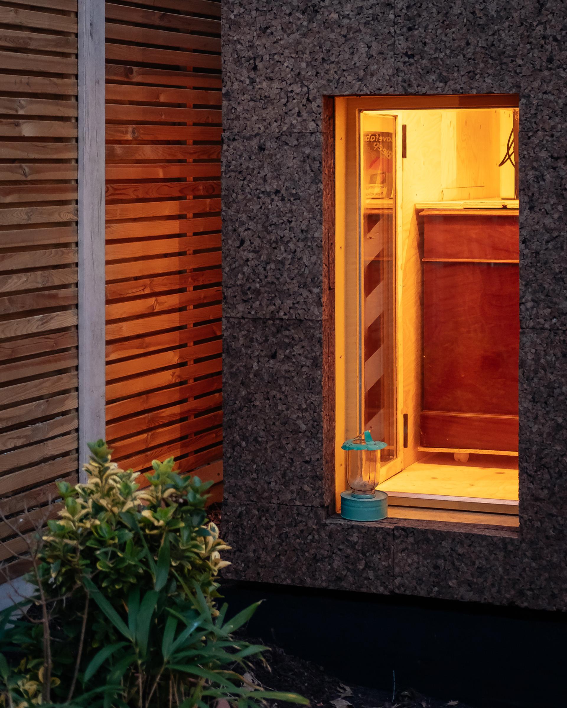 A warmly lit timber interior is visible through a window in a textured dark wall; a vintage lantern sits on the sill, whilst a leafy plant grows outside beside wooden slats—showcasing sustainable design and modular construction elements.