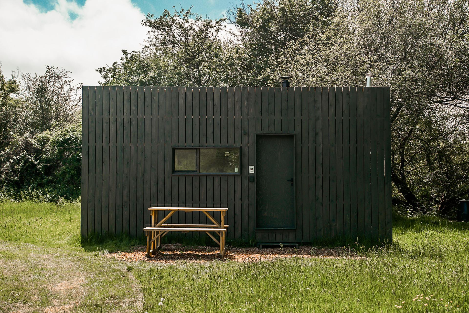 A small, dark wooden cabin with a rectangular window and door sits in a grassy field. Embracing sustainability, it features an upcycled wooden picnic table outside on gravel, surrounded by trees and greenery.