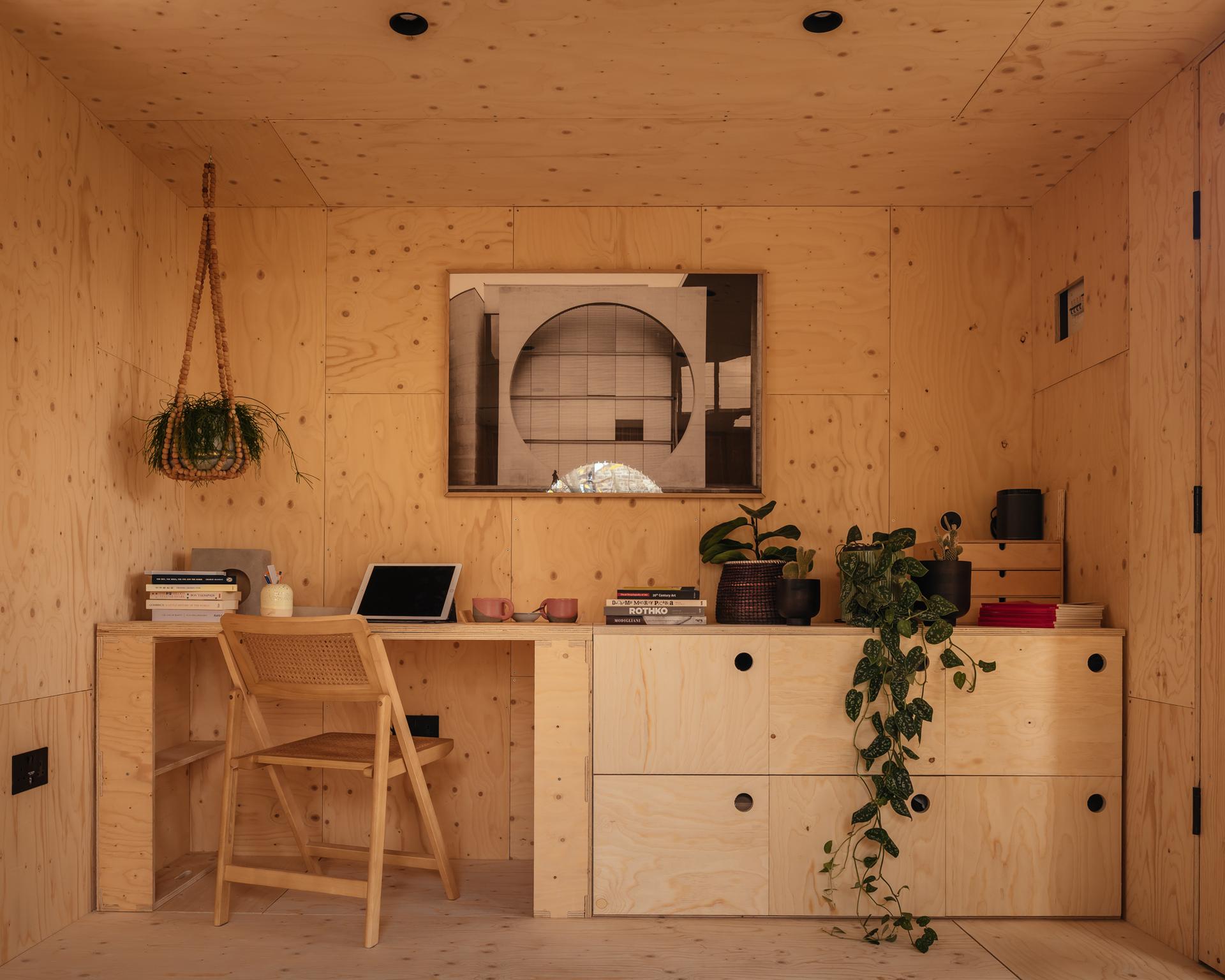 A cosy home office with light birch wood walls and furniture, featuring a modular desk with a laptop, books, potted plants, a hanging plant, a drawer unit, and a framed black-and-white geometric photo on the wall.