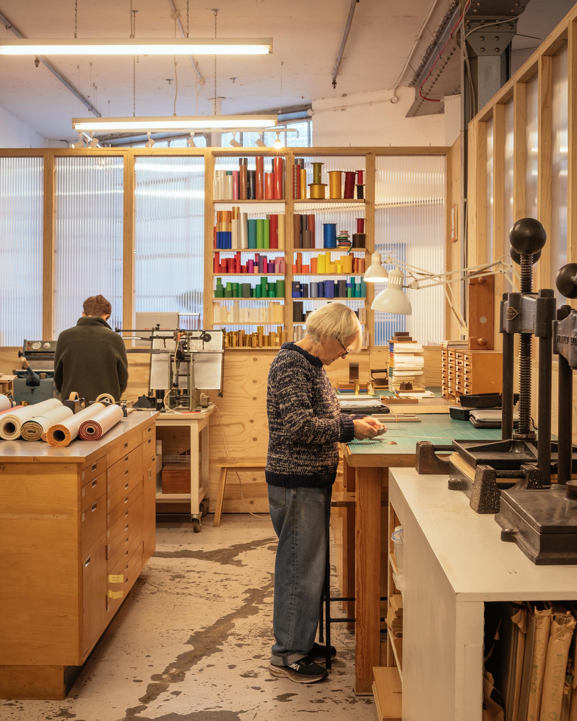 Two people work in a well-lit, organised workshop with modular wooden furniture. One stands at a table handling materials, while the other works in the background. Colourful books are neatly arranged on a shelf against the back wall.