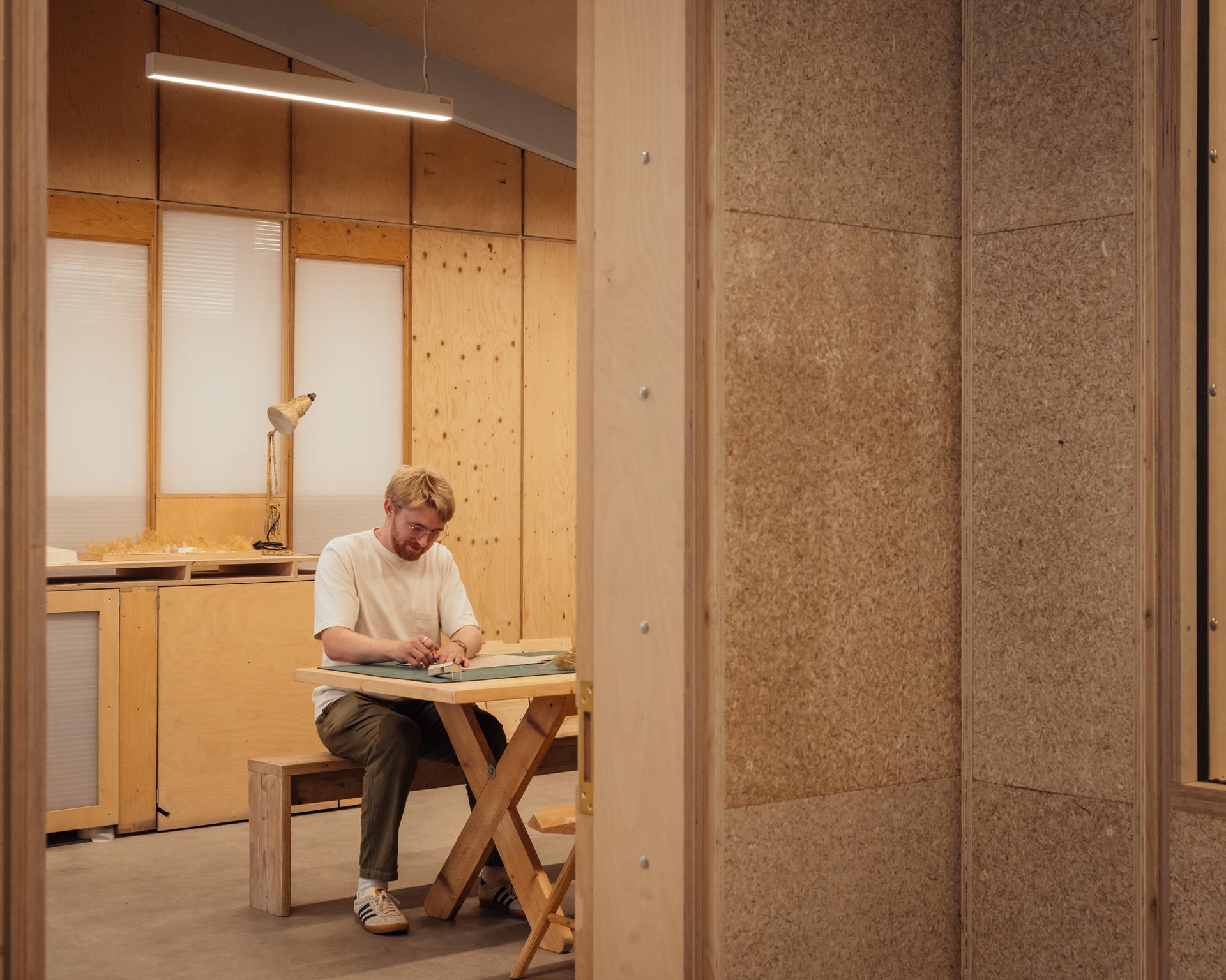 A person sits alone at a wooden table in a modern, minimalist room at Vyner Studios, working with papers. Warm lighting and wooden walls create a cosy atmosphere.