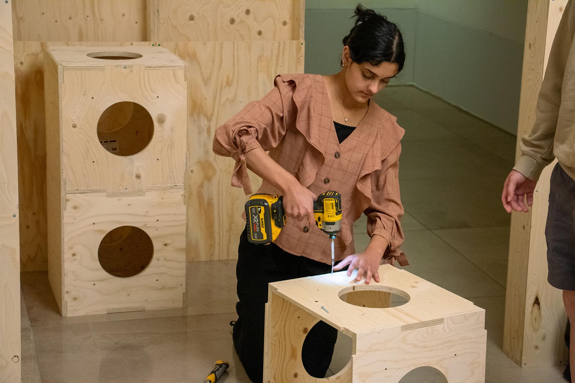 A person kneels on the floor, using a power drill to assemble a sustainable plywood box with circular cut-outs. More wooden boxes and tools are nearby, and another person stands partially visible on the right.