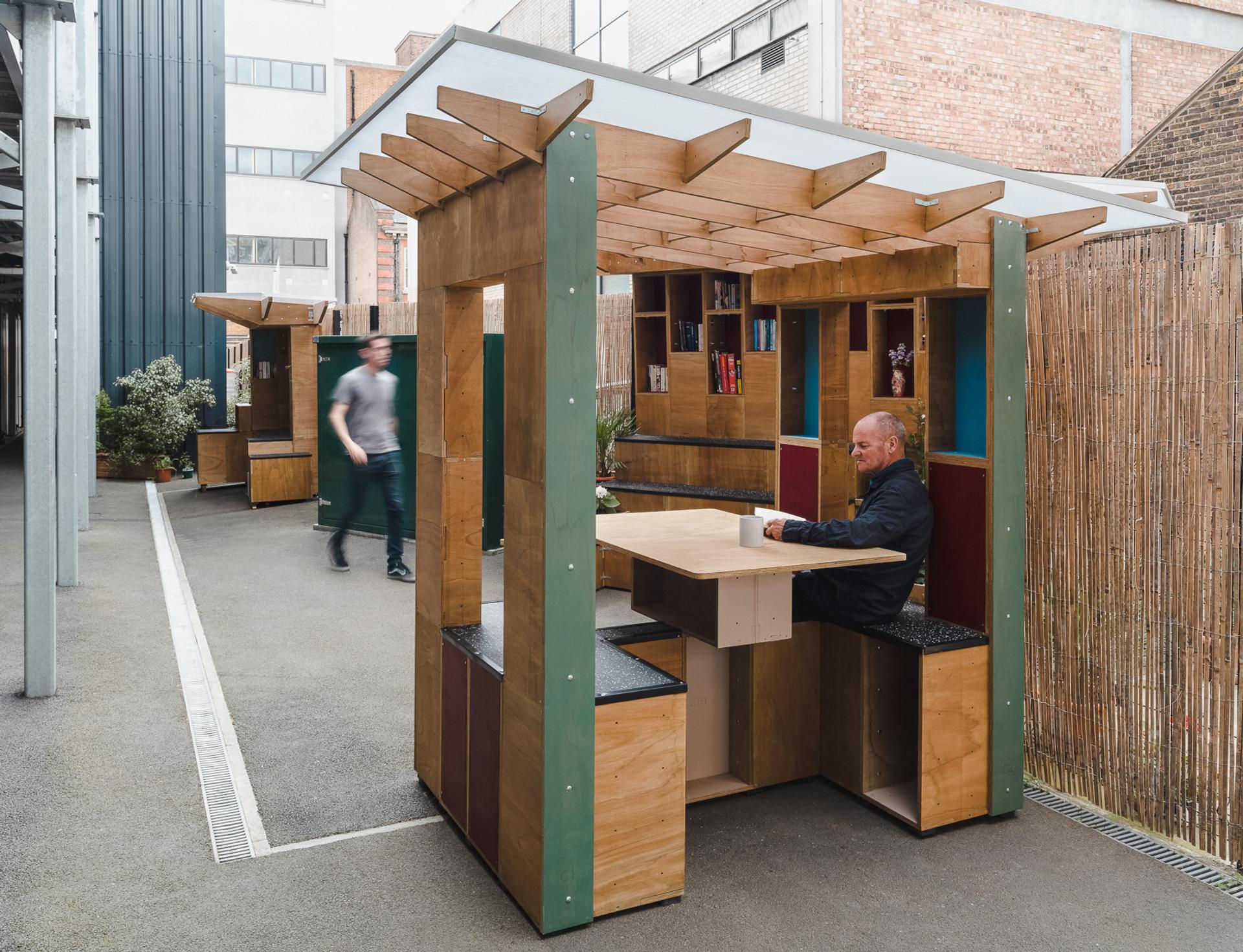 A man sits alone at a wooden outdoor stall with a roof, drinking from a cup—perhaps embracing reuse. Another person walks by in the background. The stalls are set along a paved area with wooden fencing and urban buildings nearby.