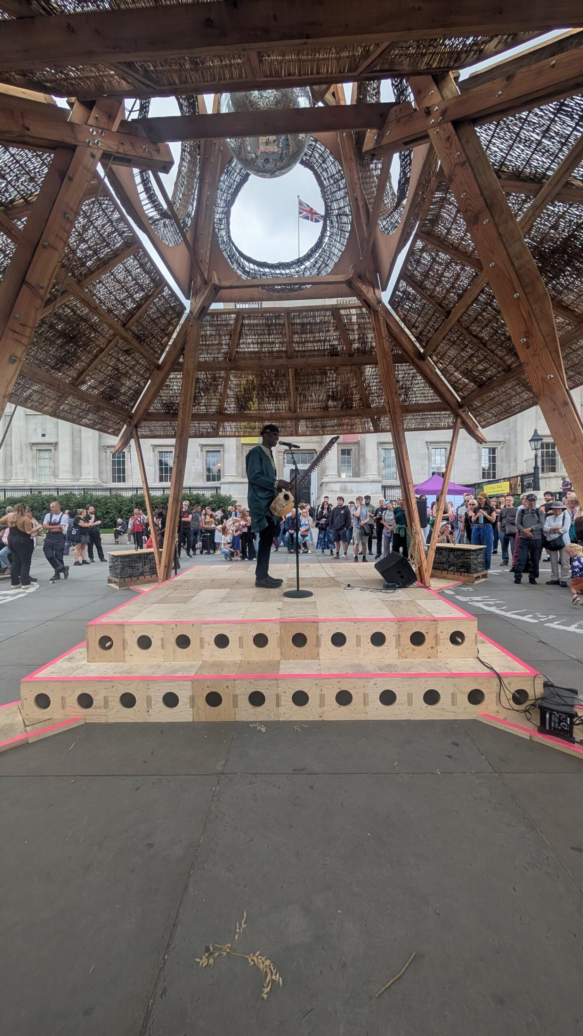 A musician plays guitar and sings on a wooden stage with a geometric roof—crafted from upcycled materials—surrounded by an audience in an outdoor urban setting. Buildings and people are visible in the background.